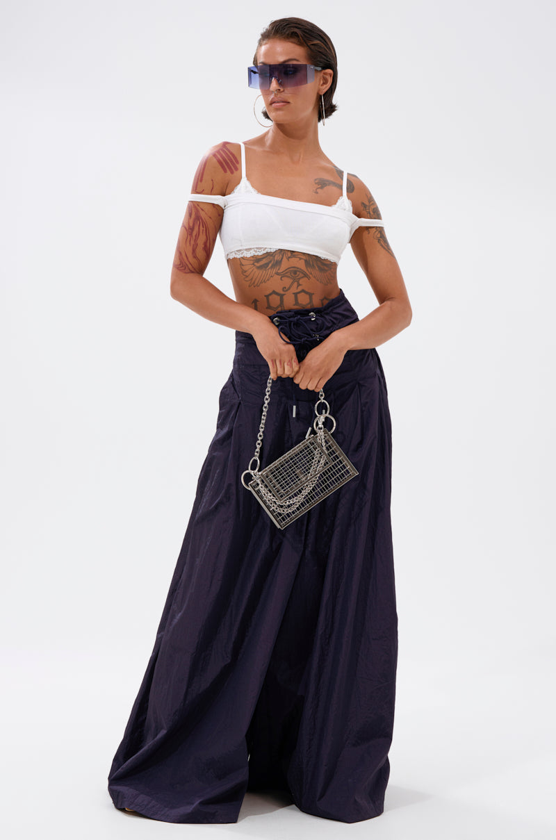 A tattooed woman wears a white crop top with cut-out shoulders, large sunglasses, and holds a small metallic handbag. She stands against a plain white background in DANCE THE NIGHT AWAY DENIM PALAZZO PANT with a wide-leg silhouette.