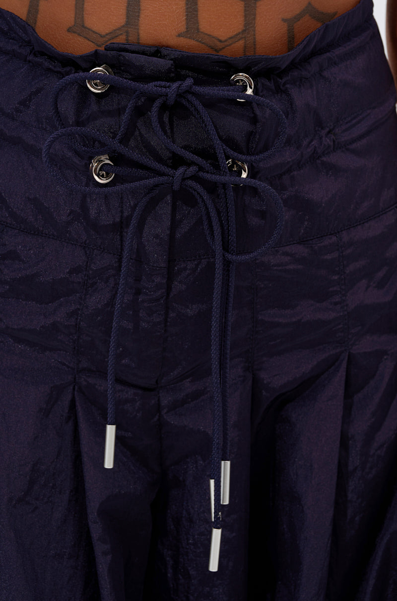 Close-up of the DANCE THE NIGHT AWAY DENIM PALAZZO PANT in dark blue with a shiny finish, lace-up drawstring waistband with silver metal tips, and high-rise fit. A geometric tattoo is partially visible above the waistband.