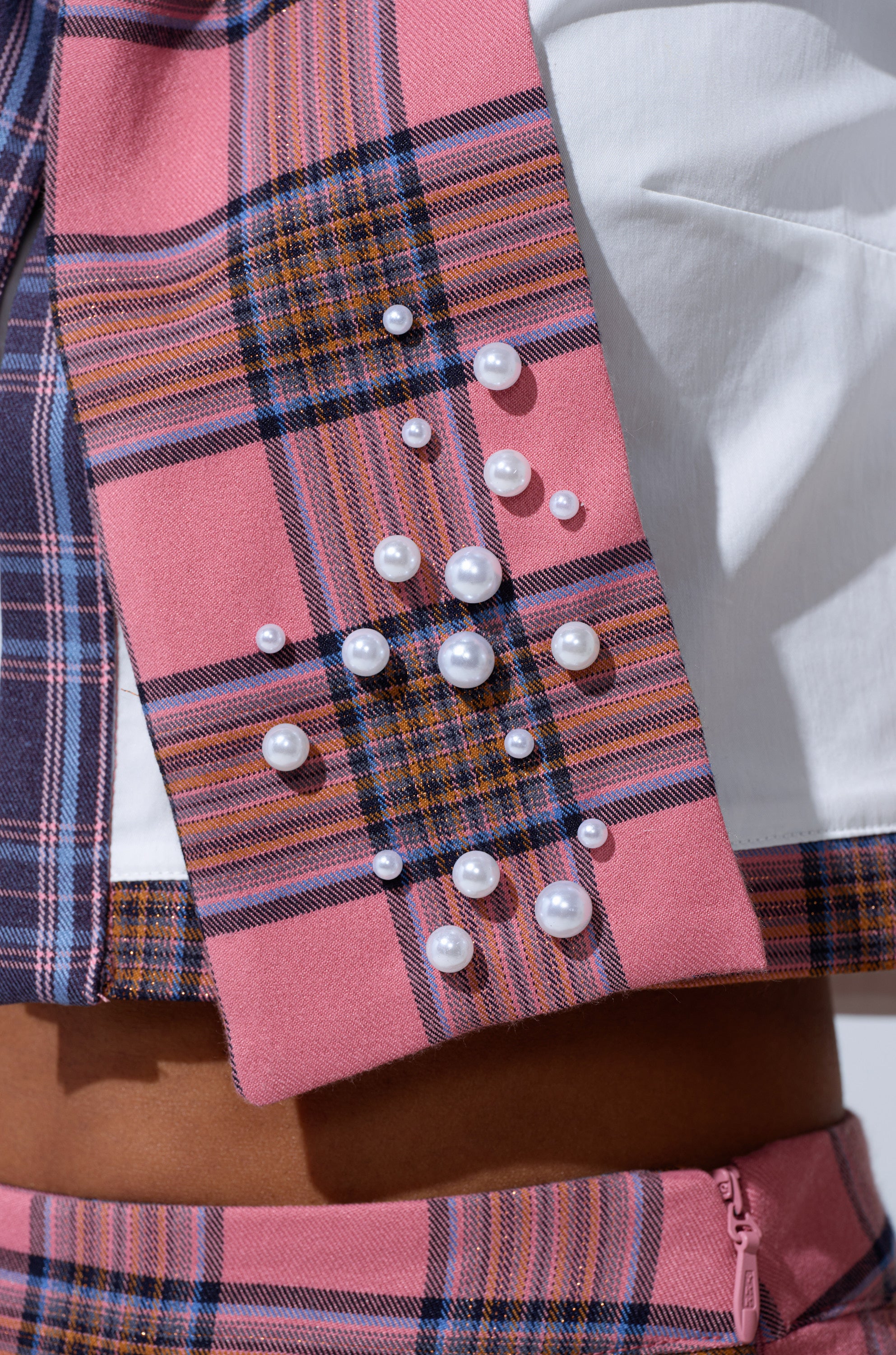 Close-up of the DREKE OVER THE TOP BOW NECKLACE, featuring pearl embellishments on a pink and blue plaid background, paired with a white top and a subtle glimpse of bare skin below.