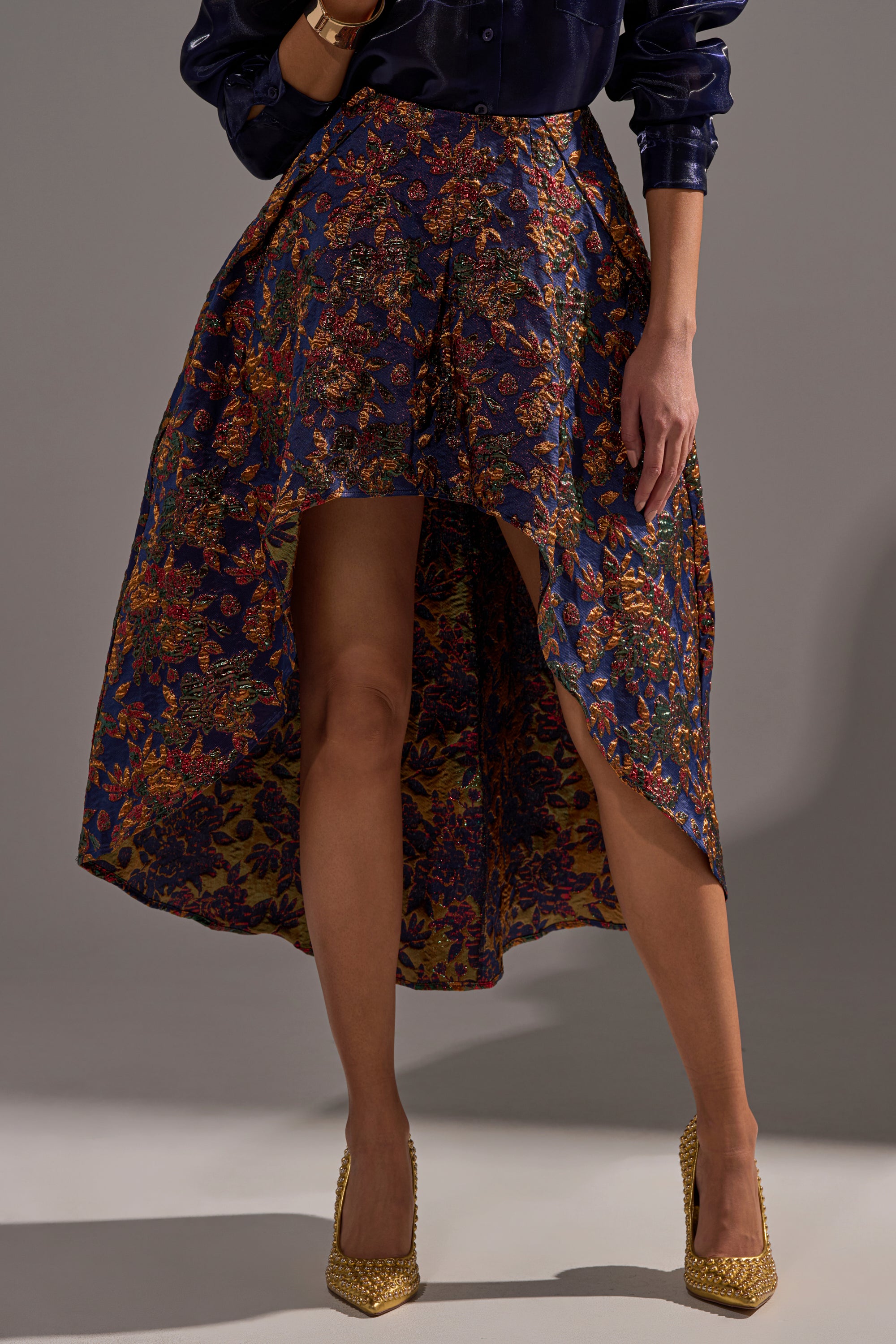 Wearing the STAR OF THE NIGHT BROCADE MIDI SKIRT and a shiny dark blue blouse with gold pointed-toe heels, a person poses against a neutral gray background.