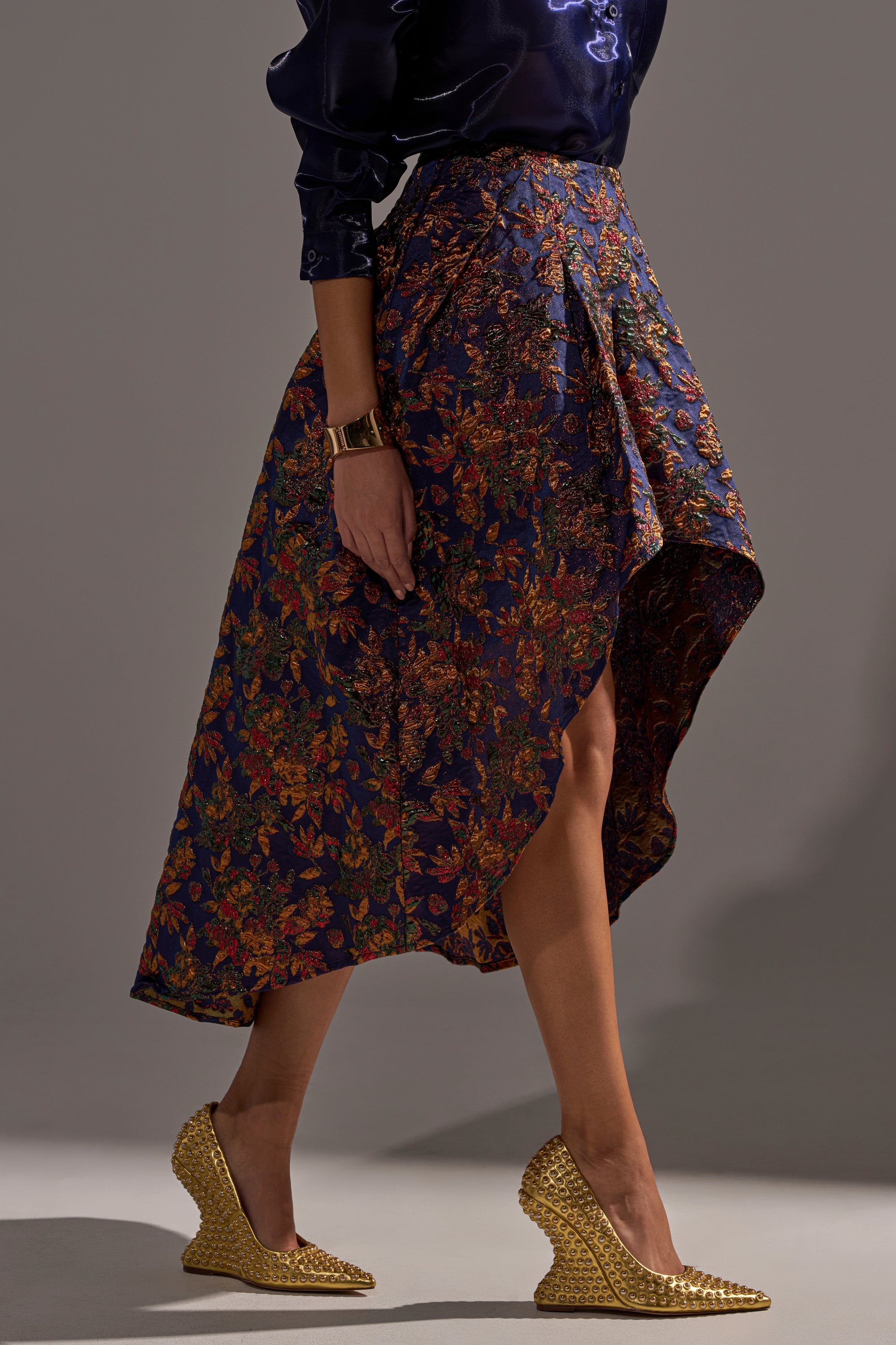 Someone is wearing the STAR OF THE NIGHT BROCADE MIDI SKIRT with metallic detail, paired with a dark satin blouse, gold bracelet, and gold textured wedge heels, walking against a plain background.