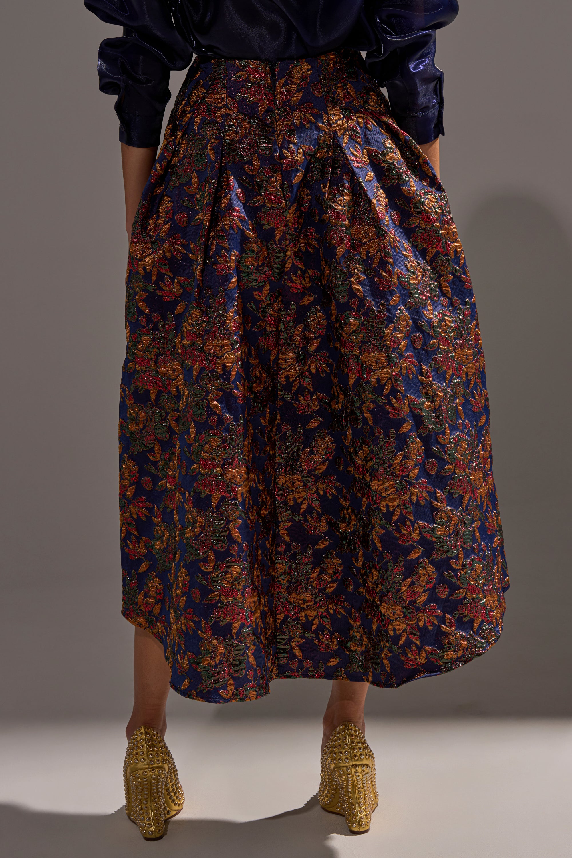A person wears the STAR OF THE NIGHT BROCADE MIDI SKIRT, a high-waisted, ankle-length dark blue skirt with a colorful floral pattern, paired with textured gold shoes. The upper body is mostly cropped out of the image.