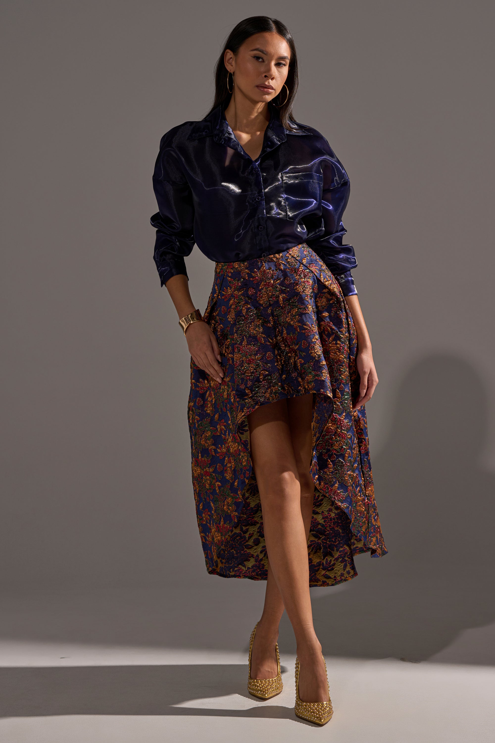 A woman poses against a plain background wearing a shiny navy button-up shirt and the STAR OF THE NIGHT BROCADE MIDI SKIRT in purples and oranges, paired with gold heels, metallic accents, and a gold bracelet.