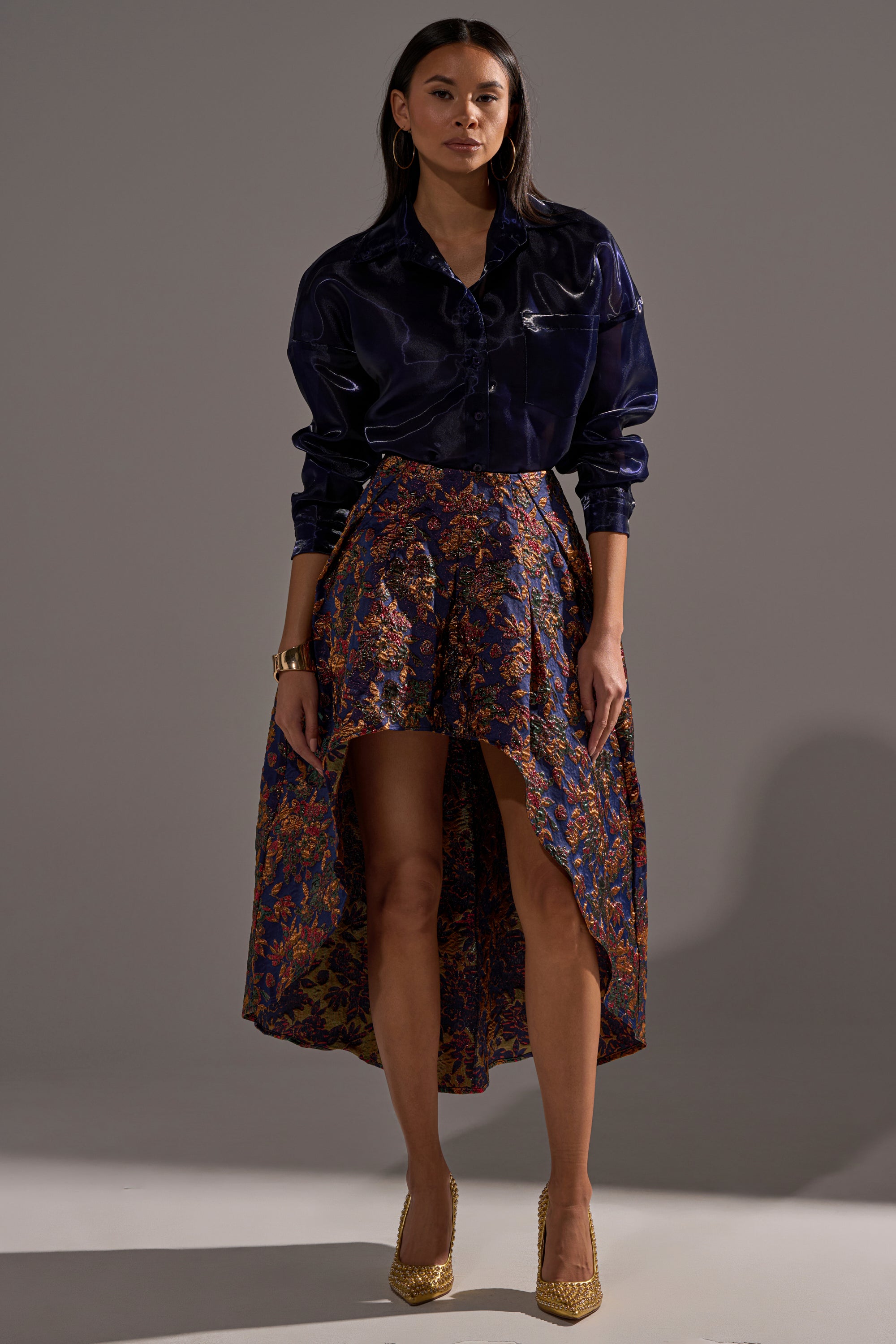 A woman poses against a plain gray backdrop wearing the STAR OF THE NIGHT BROCADE MIDI SKIRT, a shiny dark blue blouse, gold heels, hoop earrings, a bracelet, and has straight dark hair.