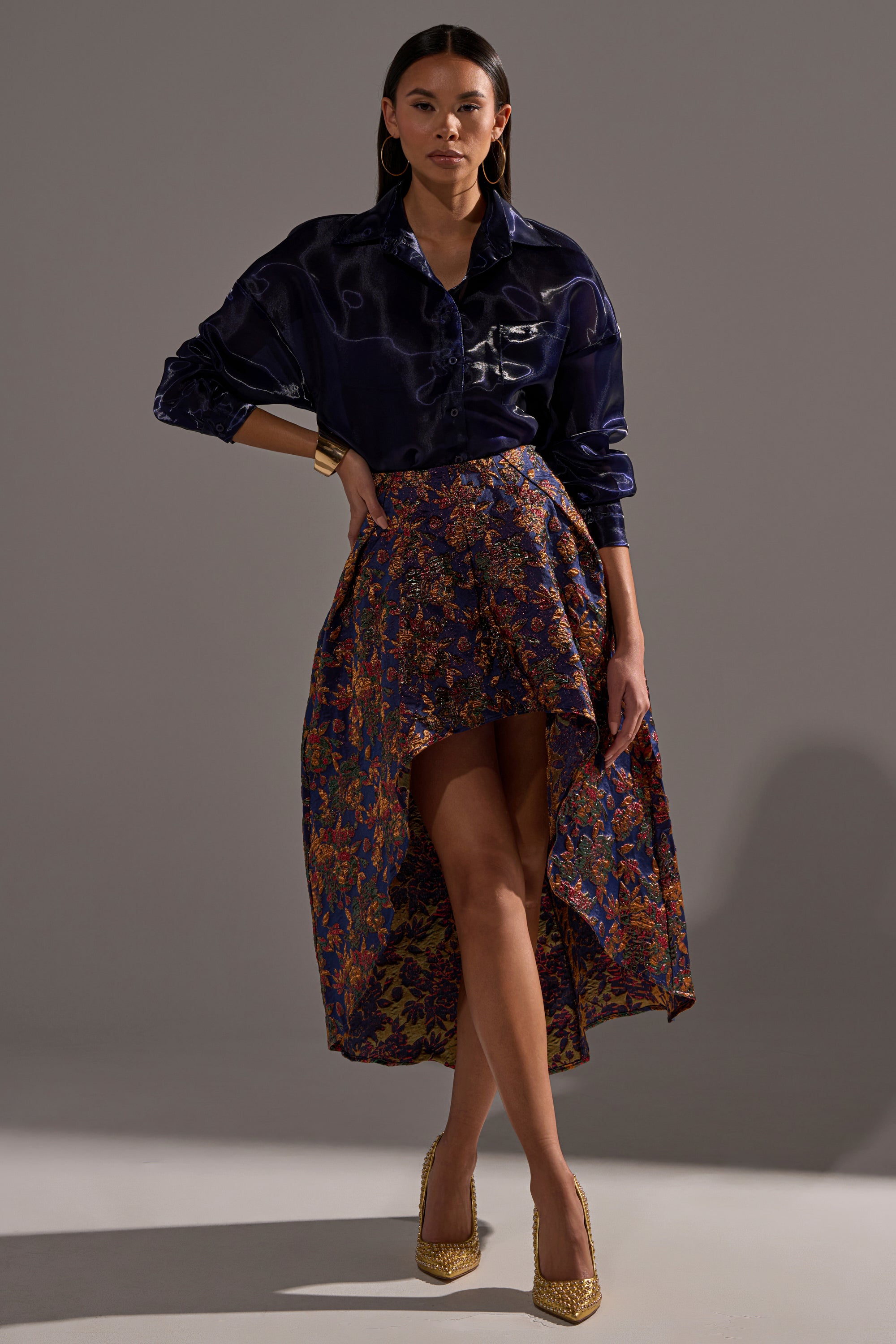 A woman stands confidently in the STAR OF THE NIGHT BROCADE MIDI SKIRT, paired with a dark satin shirt, gold hoop earrings, a bangle, and pointed gold heels. The softly lit, plain backdrop keeps the focus on her chic ensemble.