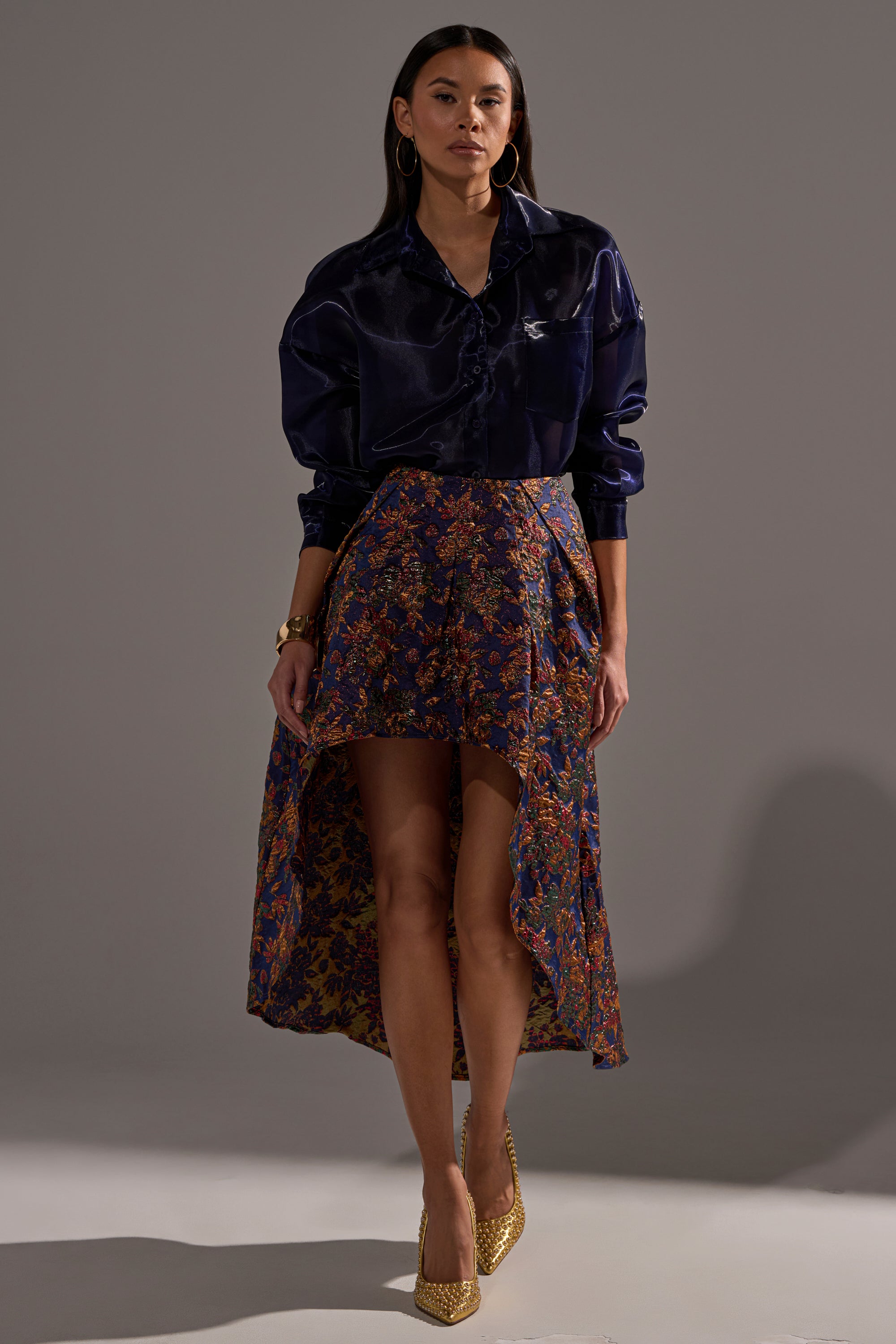 A woman poses against a gray background wearing the STAR OF THE NIGHT BROCADE MIDI SKIRT, featuring red and gold patterns with metallic details, paired with a dark silk blouse, gold hoop earrings, a bracelet, and gold heels.