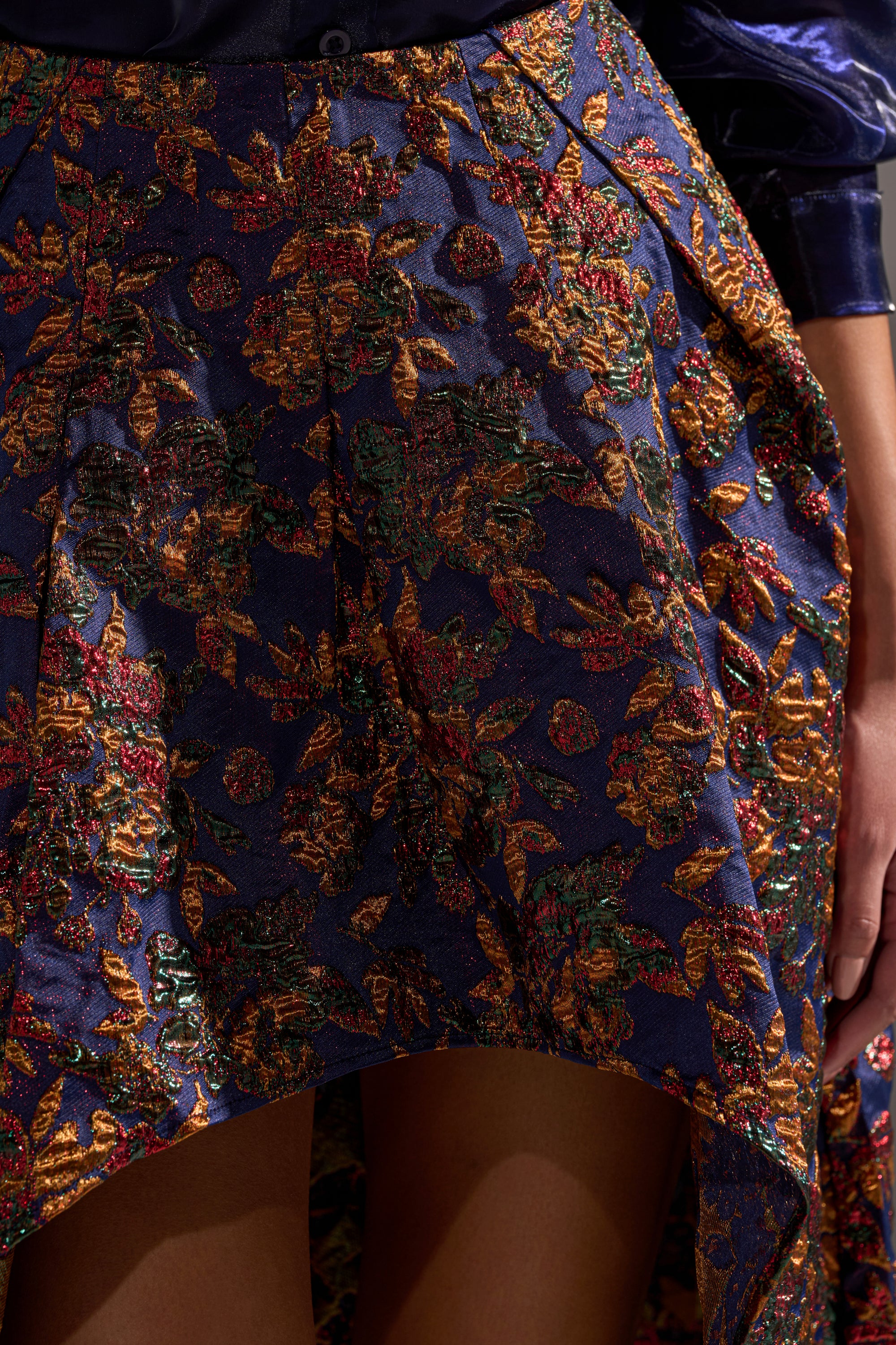 A close-up of the STAR OF THE NIGHT BROCADE MIDI SKIRT, a purple brocade midi skirt with an asymmetric hem, colorful floral pattern, and metallic detailing, worn by someone with their hand by their side.