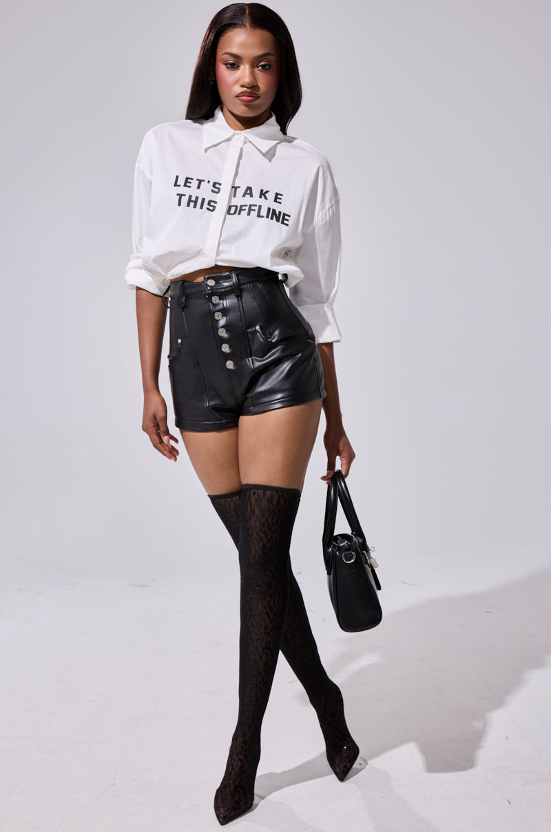 A woman poses confidently in a white "LET’S TAKE THIS OFFLINE" shirt, SKY HIGH FAUX LEATHER SHORTS, patterned thigh-high stockings, black heels, and holds a chic black handbag.
