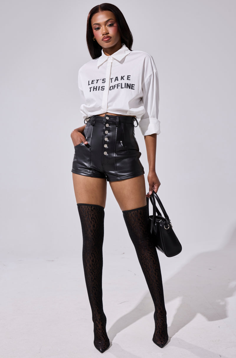A woman stands confidently in a white "LET’S TAKE THIS OFFLINE" shirt, SKY HIGH FAUX LEATHER SHORTS, black patterned thigh-high stockings, black heels, and holds a chic black handbag.