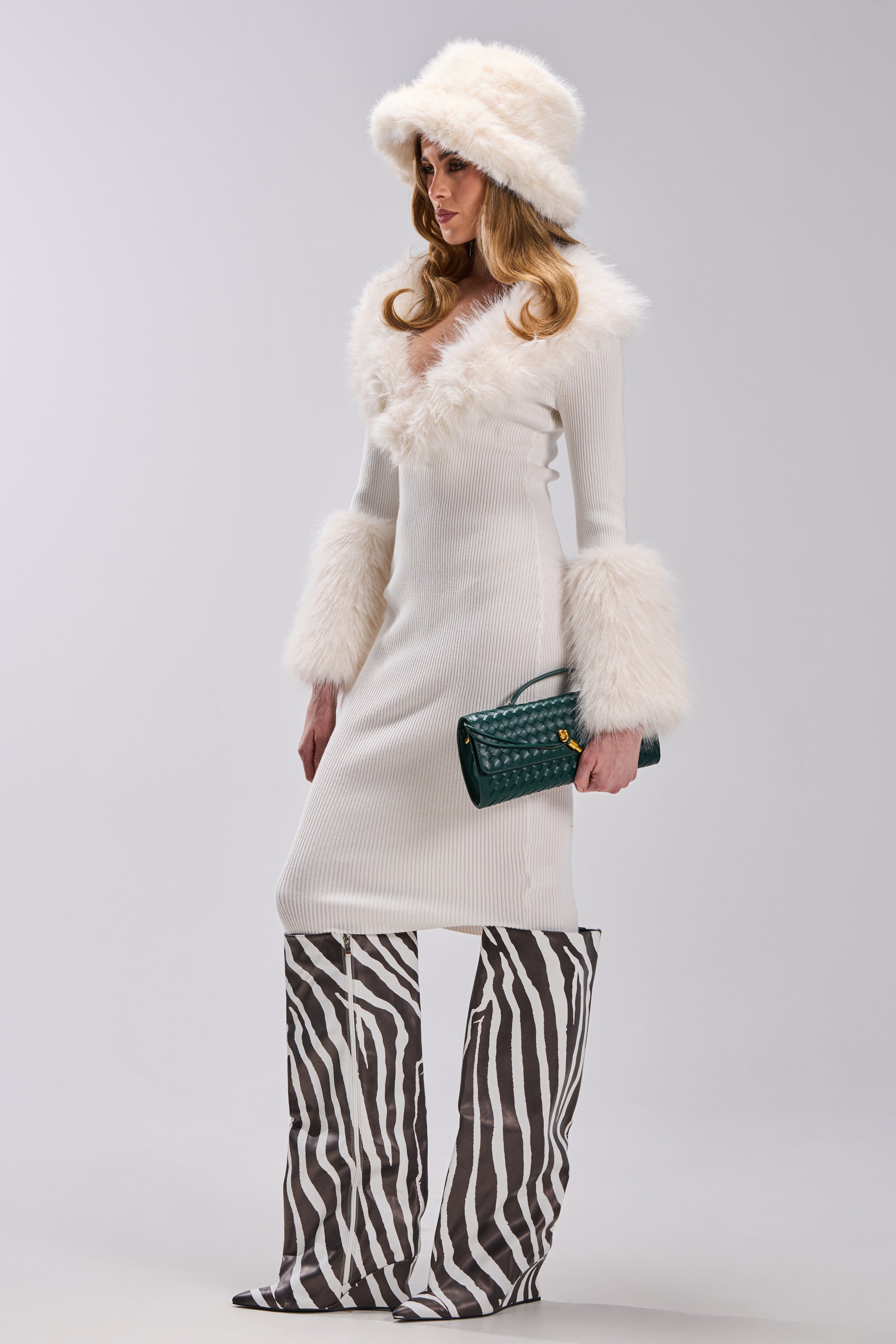 A woman wears the CELINE SWEATER MIDI DRESS WITH FUR TRIM, styled with a white furry hat, matching fur cuffs, a green handbag, and tall zebra-print boots. She stands against a plain gray background.