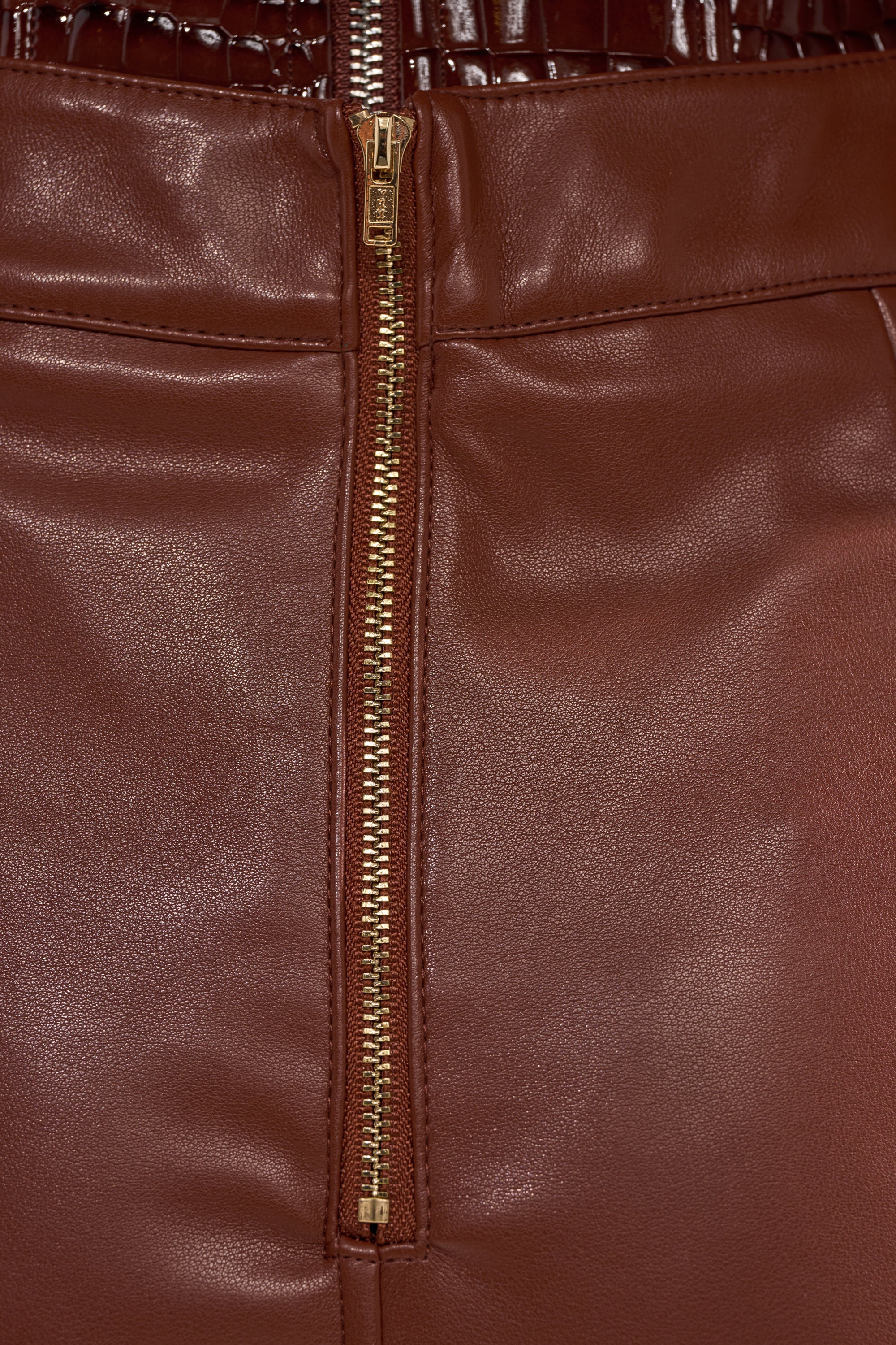 Close-up of the SOMETHING DIFFERENT FAUX LEATHER MIDI SKIRT in brown, featuring a central gold zipper. The smooth faux leather and detailed seam stitching showcase its refined craftsmanship.