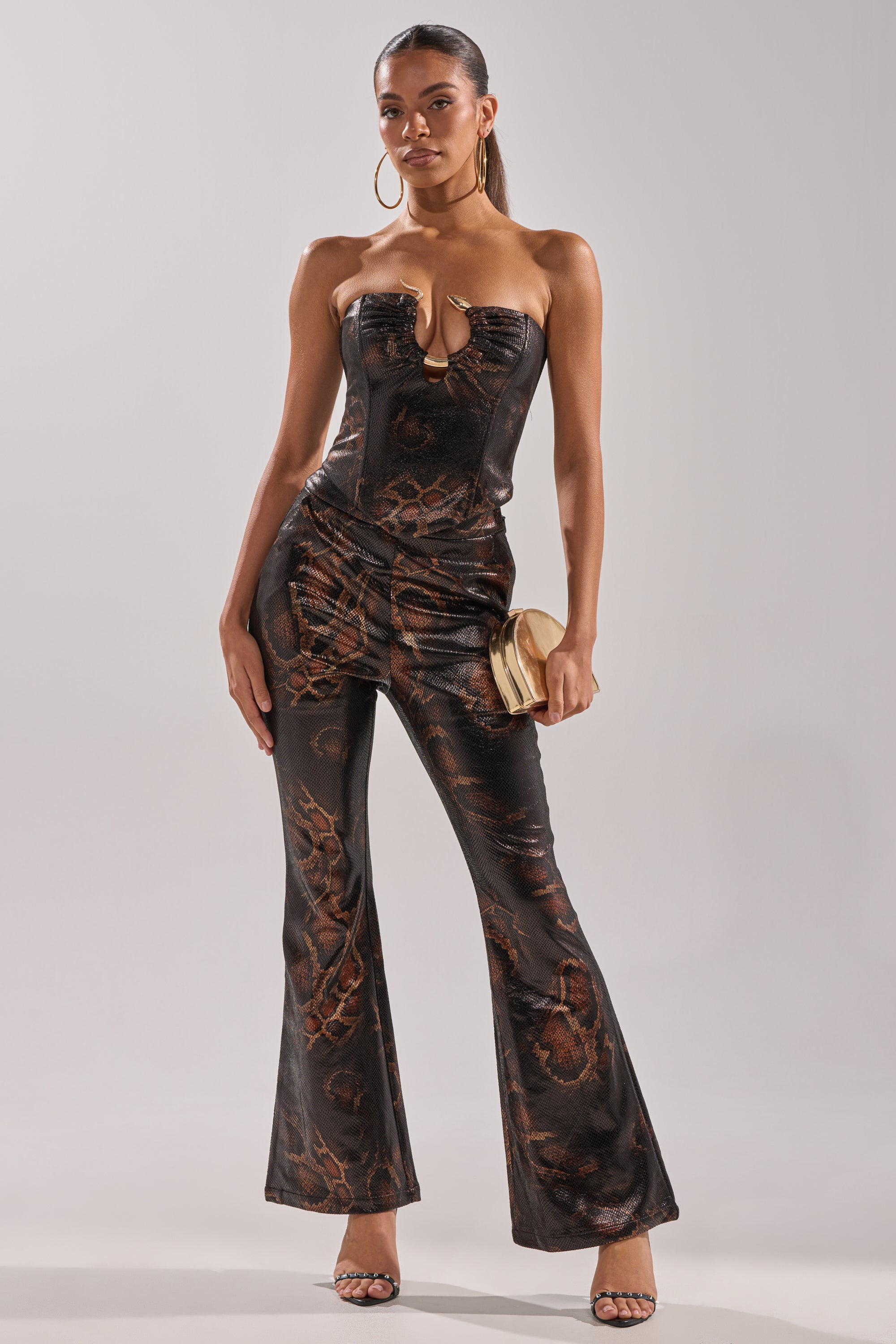 Woman stands posing in the BEING BAD SNAKE TROUSER, featuring a black and brown snakeskin pattern. She holds a beige clutch, wears hoop earrings and heeled sandals, with her hair pulled back.