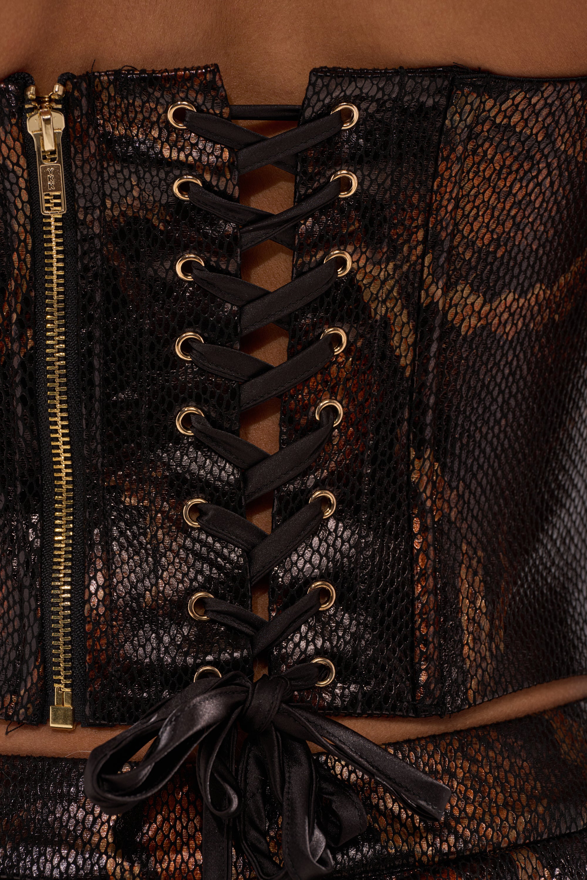 Close-up of the BEING BAD SNAKE CORSET in faux snakeskin with gold eyelets, gold snake hardware, black lace-up ribbon, and a gold left-side zipper, worn by a person with medium brown skin.