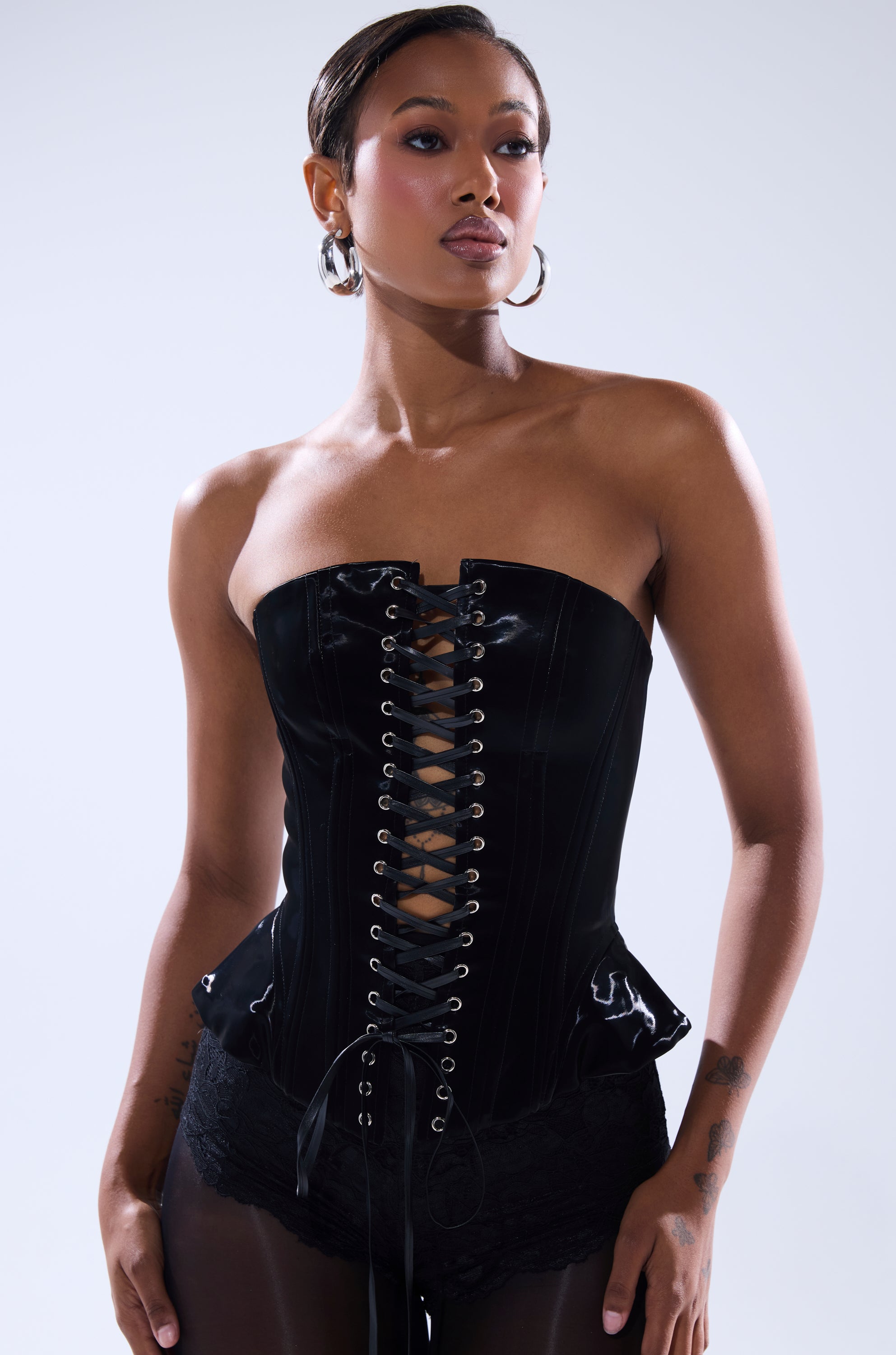 A woman poses confidently in the SMOOTH TRANSITION LACE UP CORSET, featuring a faux patent leather finish and paired with black lace bottoms. Her bold style is highlighted by slicked-back hair, hoop earrings, and visible tattoos.