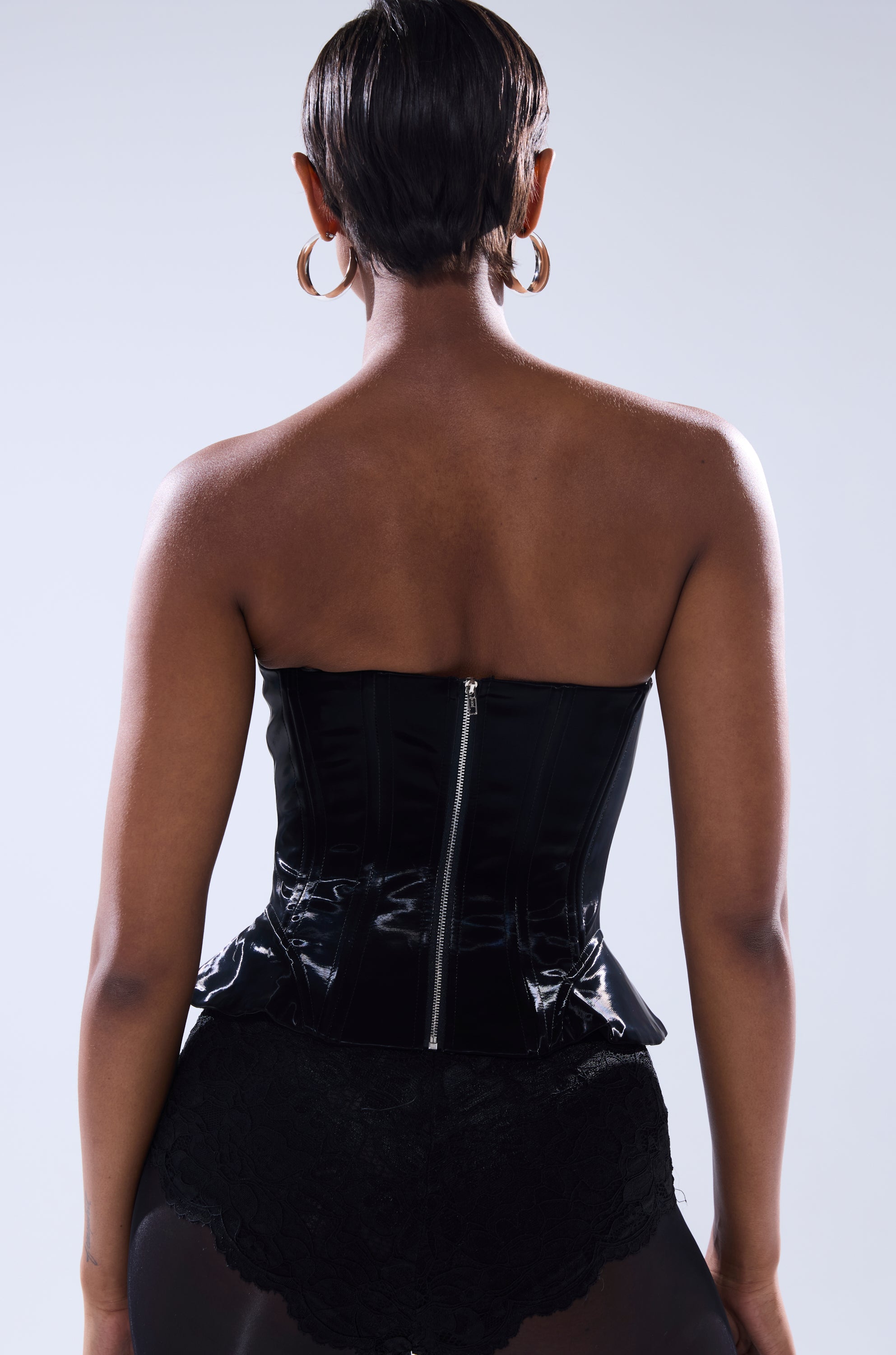 A person with short hair, large hoop earrings, and black lace bottoms is shown from behind against a plain light background, wearing the SMOOTH TRANSITION LACE UP CORSET—a faux patent leather top with a zipper down the back.