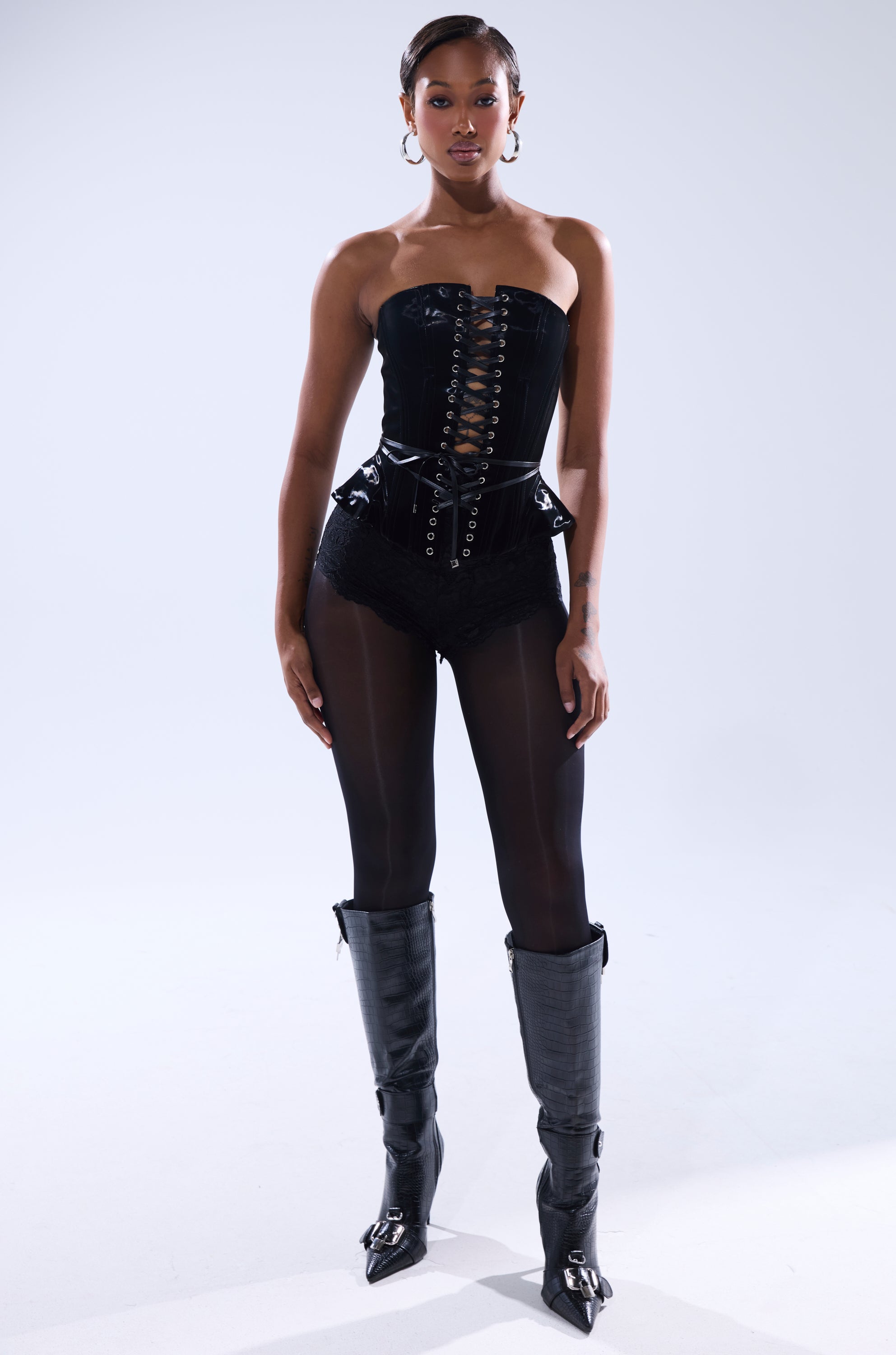 A woman stands confidently against a light background, wearing the SMOOTH TRANSITION LACE UP CORSET with high-waisted shorts, sheer tights, knee-high boots, hoop earrings, and neatly styled hair.
