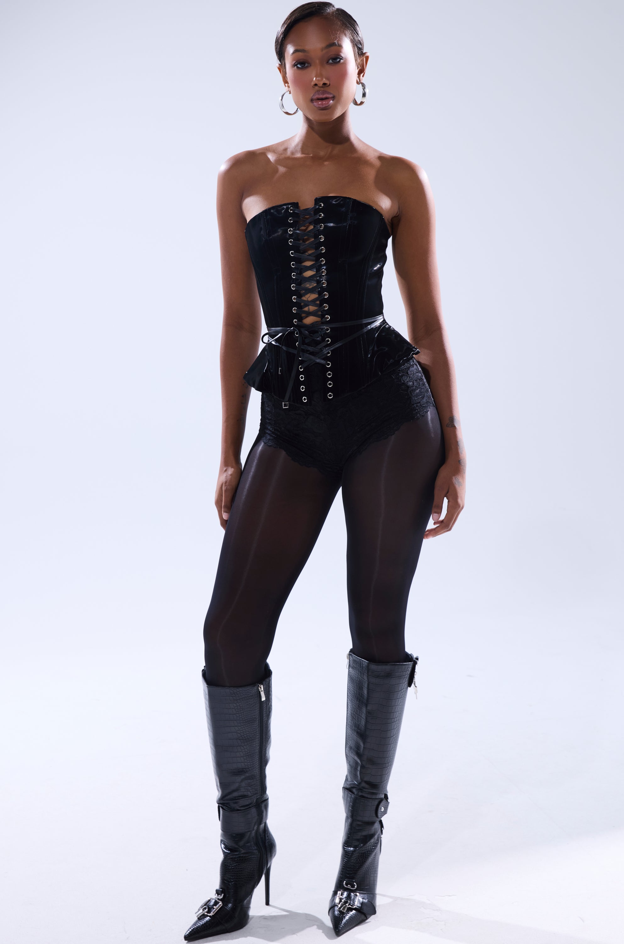 A woman poses confidently in the SMOOTH TRANSITION LACE UP CORSET, paired with high-waisted shorts, sheer black tights, and knee-high boots against a light gray background. She accessorizes with hoop earrings and a sleek hairstyle.