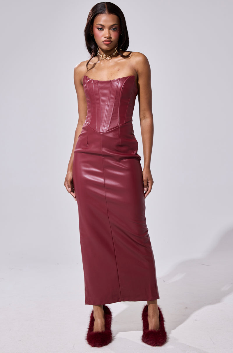 A woman models the JULIETTE PU MIDI DRESS IN BURGUNDY, a strapless, fitted faux leather dress with a corset bodice and long skirt, styled with matching burgundy fuzzy heels and gold jewelry in a studio setting.