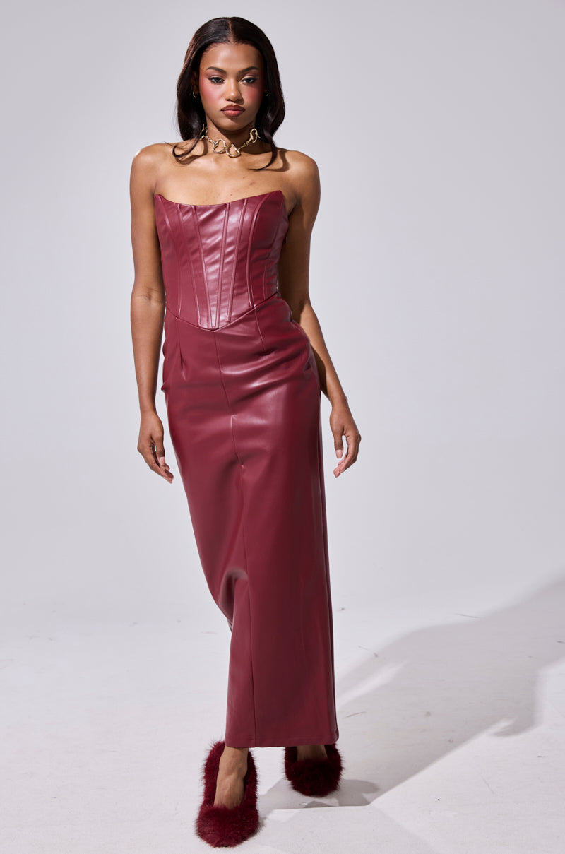 A woman stands in a studio wearing the JULIETTE PU MIDI DRESS IN BURGUNDY—a strapless, fitted faux leather midi with a structured bodice and long skirt—styled with loose hair, statement earrings, and burgundy fuzzy shoes.