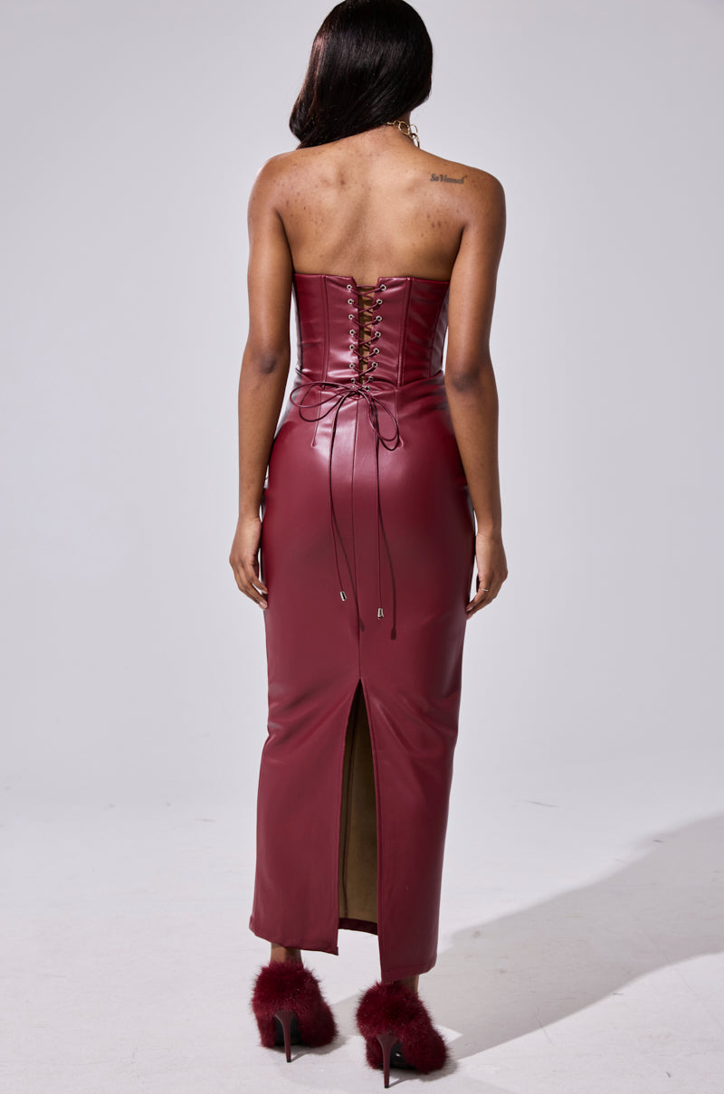 A woman wears the JULIETTE PU MIDI DRESS IN BURGUNDY, a strapless bodycon with lace-up back and back slit. She pairs it with matching fluffy-heeled shoes, her shoulder-length hair simply styled against a plain background.
