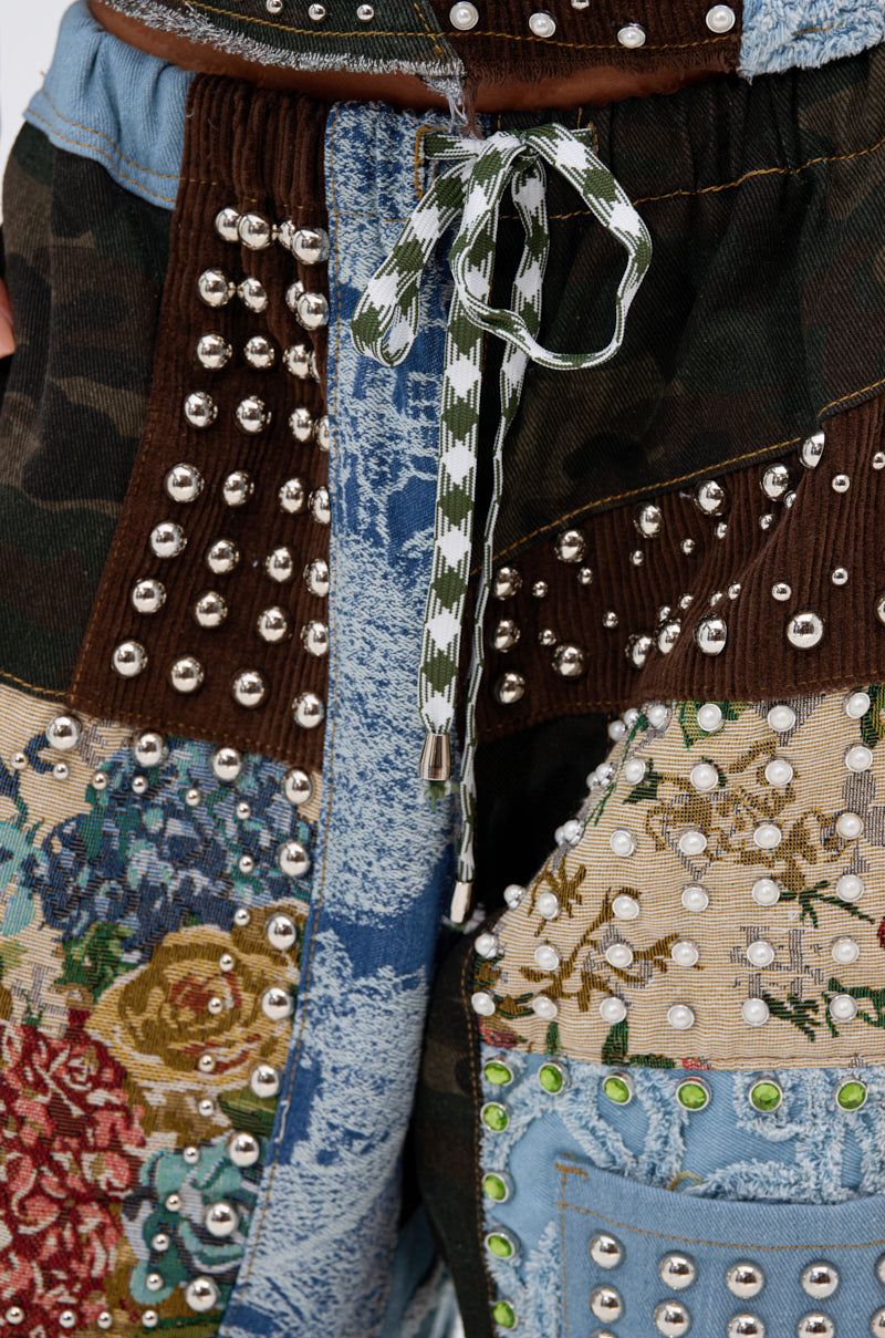 Close-up of the CALL EM CRAZY PATCHWORK JOGGER, showcasing denim, floral tapestry, and corduroy panels, detailed with metal studs, green rhinestones, and a green-and-white checkered drawstring for bold streetwear style.