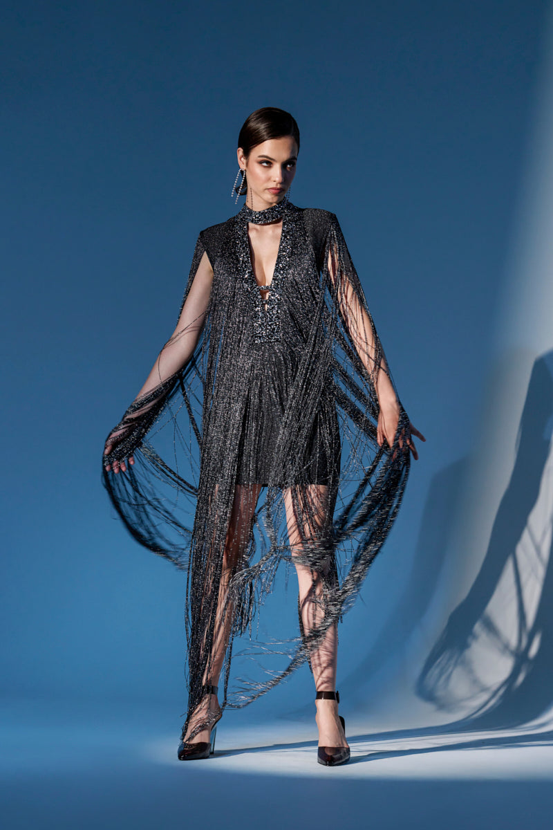 A woman poses confidently against a bold blue backdrop, wearing the MIXED MESSAGES METALLIC BANDAGE MINI DRESS WITH FRINGE. The shimmering black mini dress features dramatic fringe from her shoulders and arms, paired with high heels and statement earrings.