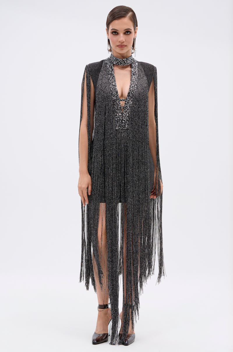 A woman poses in the MIXED MESSAGES METALLIC BANDAGE MINI DRESS WITH FRINGE, featuring a sparkly rhinestone V-neckline and long metallic fringe on the sleeves and hem. She has slicked-back hair, black heels, and stands against a white background.