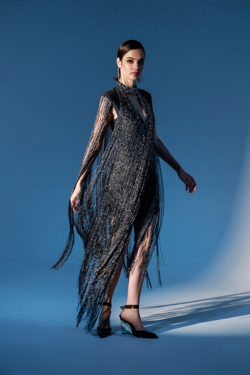 A woman poses in the MIXED MESSAGES METALLIC BANDAGE MINI DRESS WITH FRINGE, featuring a shimmering black design with a rhinestone V-neckline and sleeveless cut. She pairs it with black heels against a gradient blue backdrop and soft, dramatic lighting.
