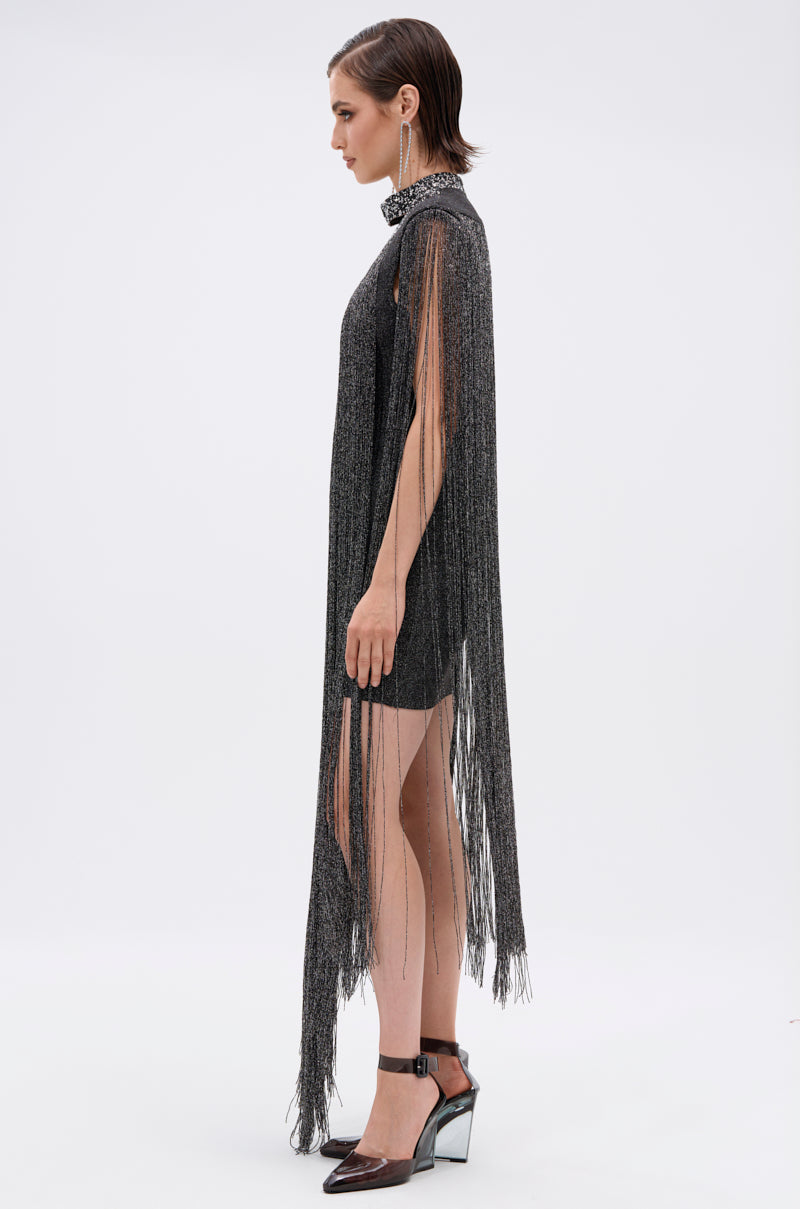 A woman with short dark hair stands in profile wearing the MIXED MESSAGES METALLIC BANDAGE MINI DRESS WITH FRINGE, featuring long silver fringe from shoulders to floor. She styles it with black heels and statement earrings against a white background.