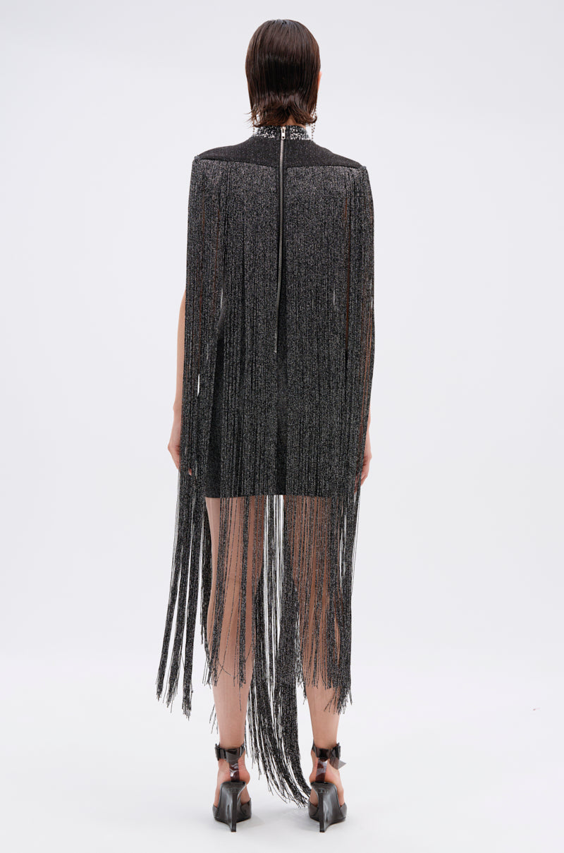 A person with short, wet-look hair faces away in the MIXED MESSAGES METALLIC BANDAGE MINI DRESS WITH FRINGE—black, with long shimmery fringe from shoulders past knees—paired with black heeled sandals against a plain white background.