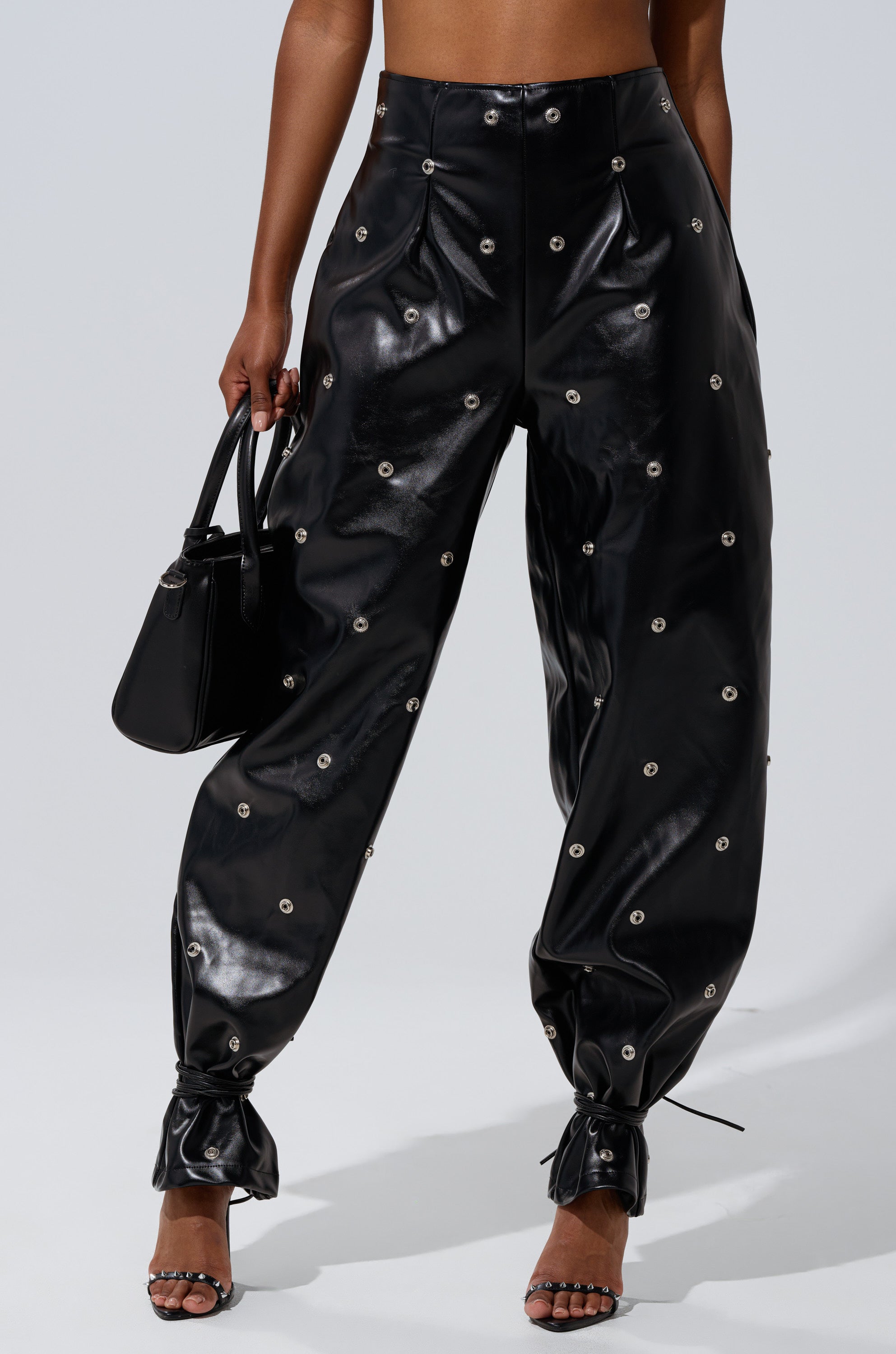 Someone wears the WORK IT BABY FAUX LEATHER BARREL PANT in shiny black with metallic grommet details, paired with a black handbag and open-toe heeled sandals. The upper body is partially visible.