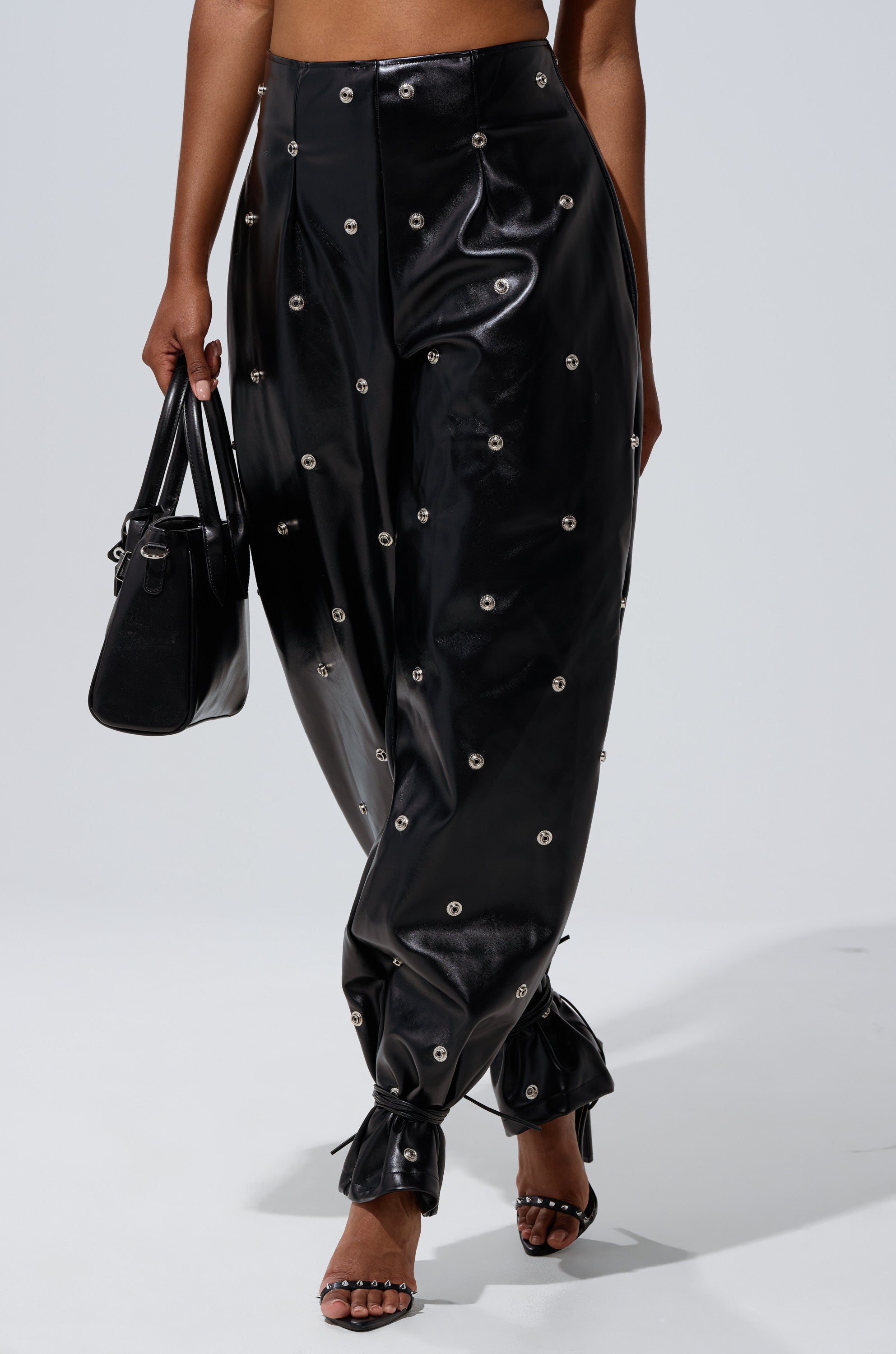 A person wears the WORK IT BABY FAUX LEATHER BARREL PANT, featuring a high-rise waist and small circular studs, paired with a black handbag and strappy black heels on a plain background.