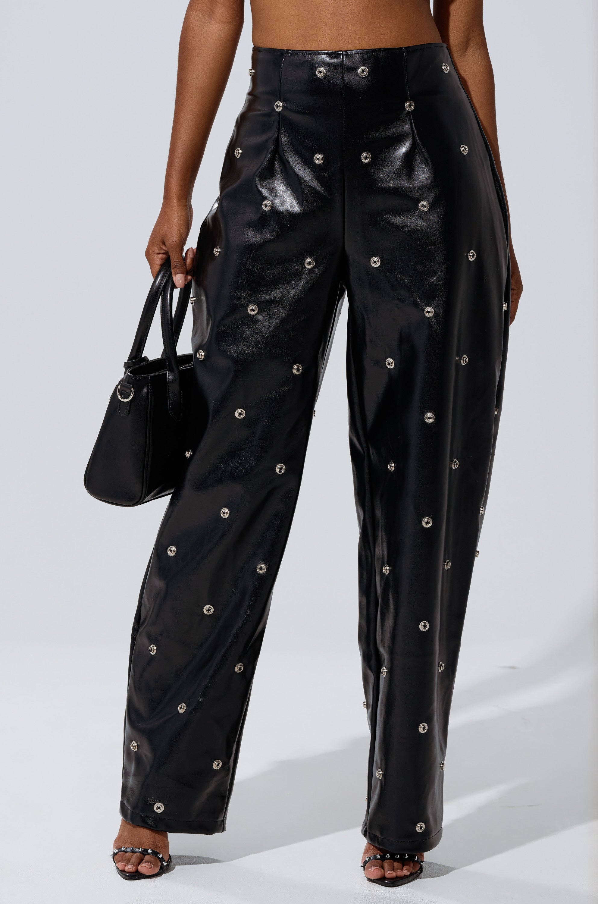 A person is shown from the waist down in WORK IT BABY FAUX LEATHER BARREL PANT, featuring a high-rise waist, black faux leather, and metallic grommet details. They hold a black handbag and wear open-toe heeled sandals; upper body and face not visible.