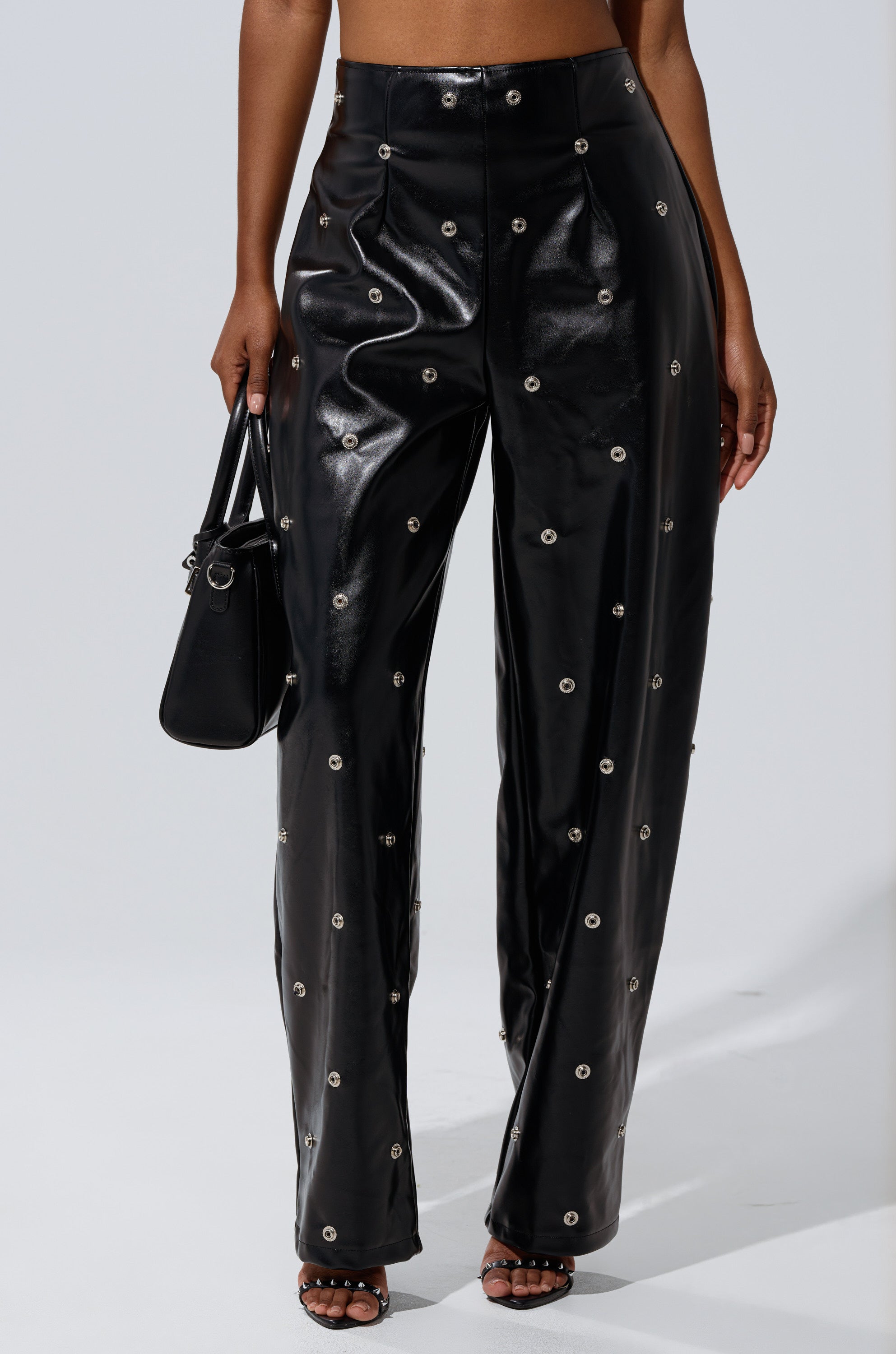 Wearing the WORK IT BABY FAUX LEATHER BARREL PANT with metallic grommet details, a person holds a small black handbag and wears black heeled sandals, shown waist-down against a plain background.