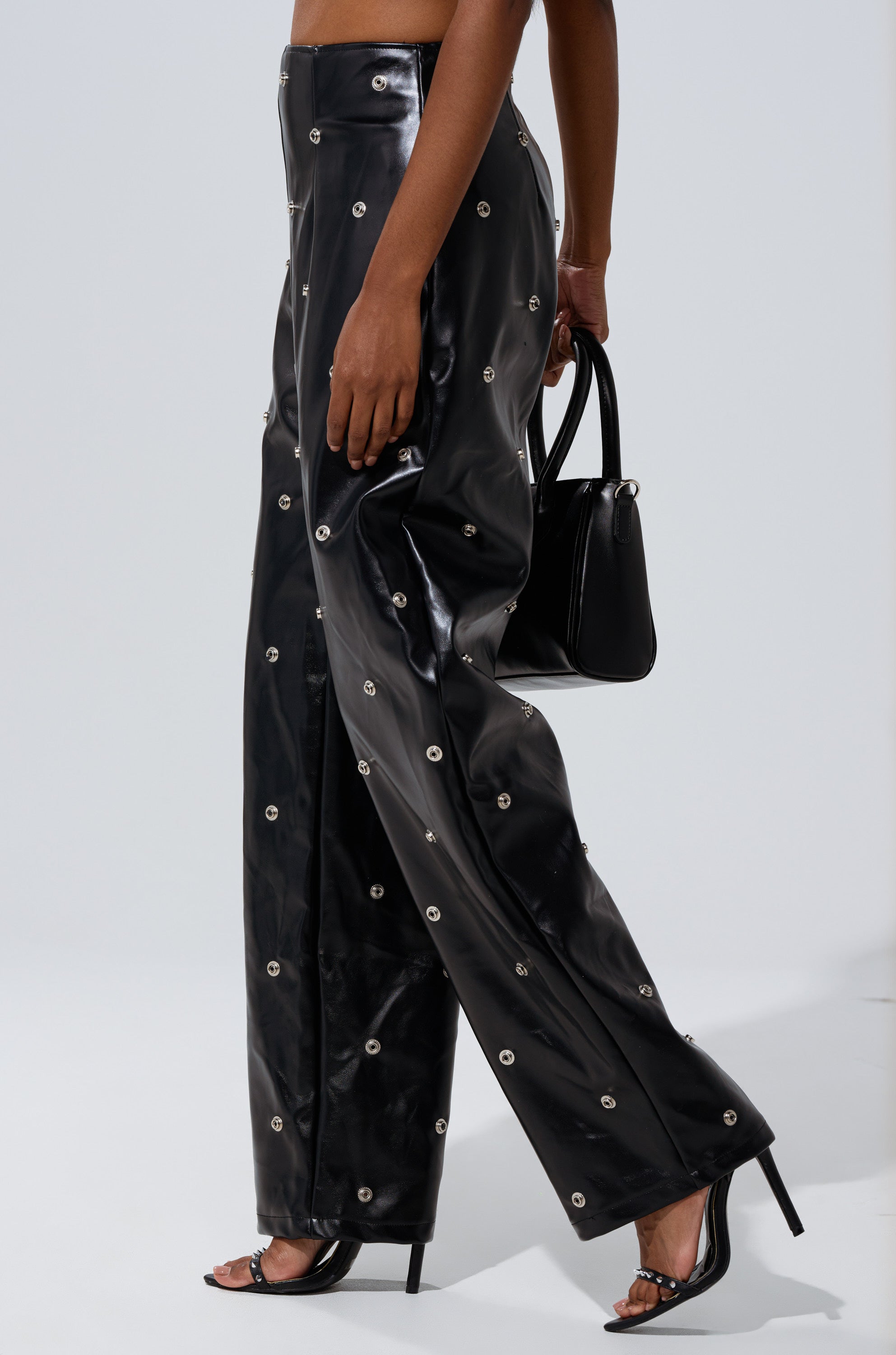 Someone wearing the WORK IT BABY FAUX LEATHER BARREL PANT with metallic grommet details, a high-rise waist, black heels, and a black handbag stands against a plain light background.