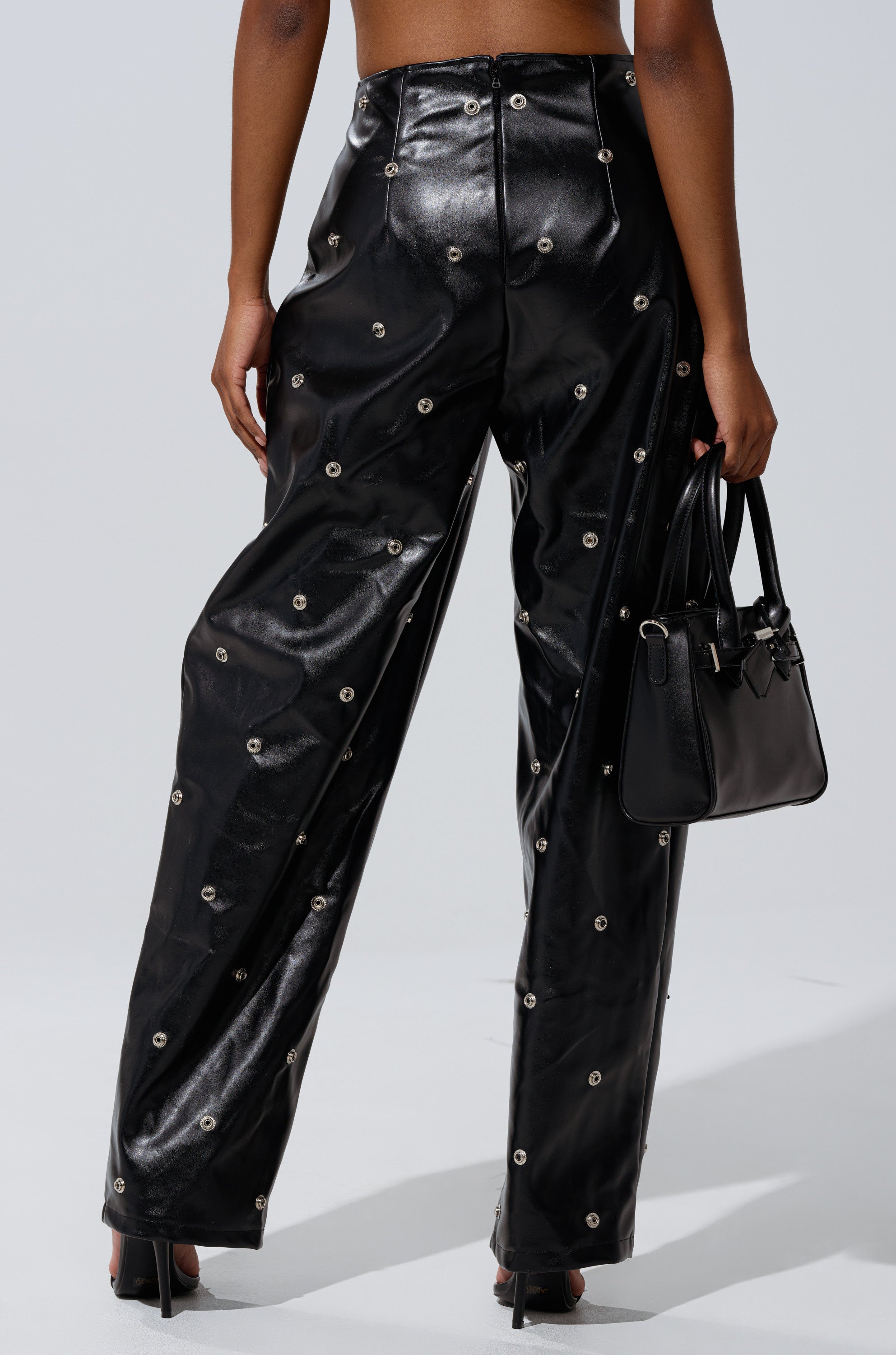 A person is seen from the back wearing the WORK IT BABY FAUX LEATHER BARREL PANT with metallic grommets, styled with heeled sandals and a black handbag.