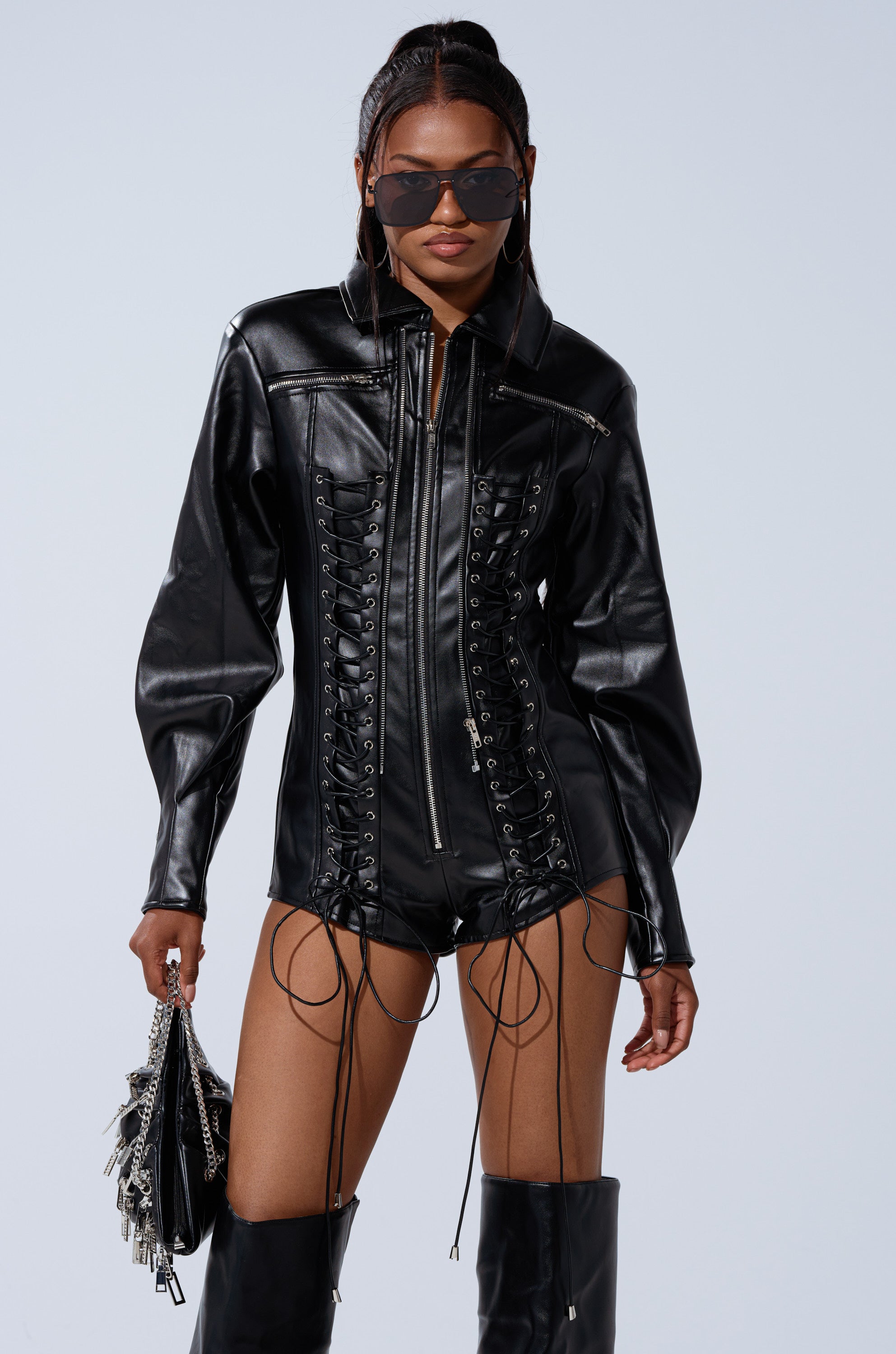 A woman poses confidently in the AT THE MAIN STAGE PU ROMPER, styled with thigh-high boots, sunglasses, and a silver-studded black handbag against a plain light background.