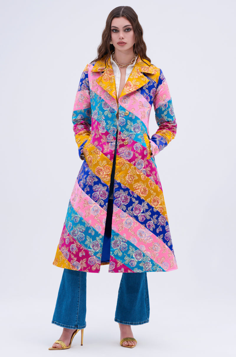 A woman faces forward against a light background, wearing the BIASED PATCHWORK BROCADE TRENCH—a long, colorful coat with diagonal floral stripes in blue, pink, yellow, and magenta—paired with blue jeans and gold heels.