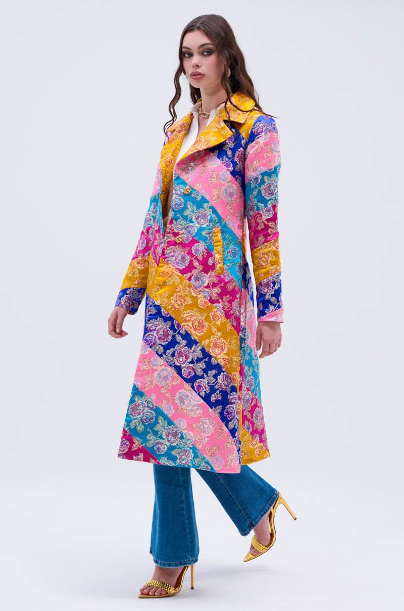 A woman models the BIASED PATCHWORK BROCADE TRENCH, a statement coat with long, colorful diagonal stripes, paired with blue jeans and gold high heels against a plain light background.