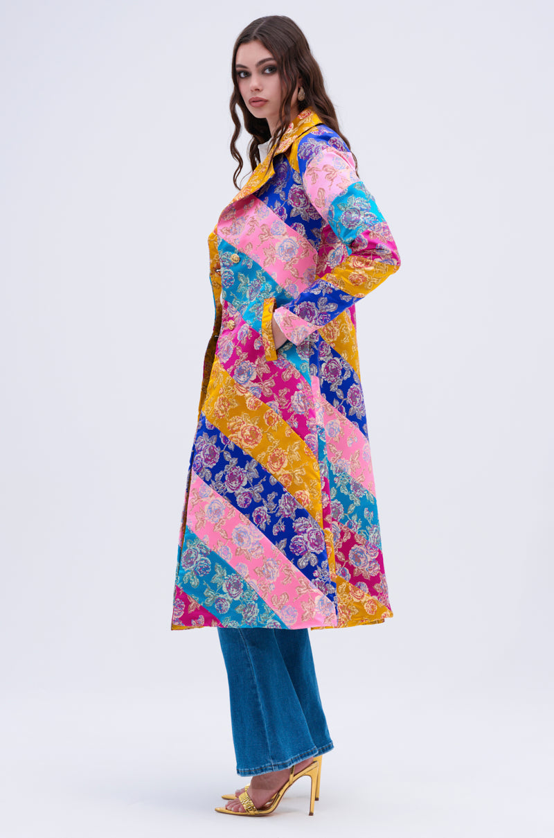 A woman stands in profile against a plain background, wearing the BIASED PATCHWORK BROCADE TRENCH—a long, colorful floral coat—paired with blue jeans and gold high-heeled sandals.