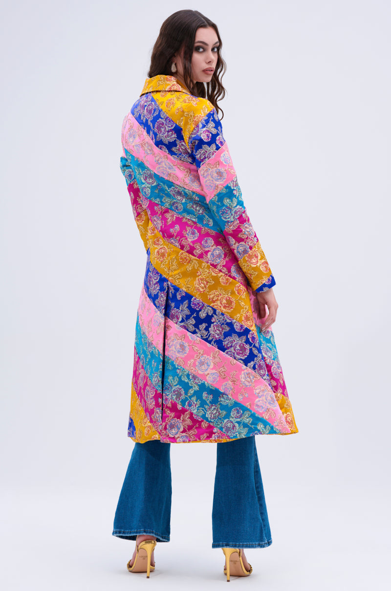 A woman faces away, wearing the BIASED PATCHWORK BROCADE TRENCH with diagonal stripes and floral designs, styled with blue flared jeans and yellow high-heeled sandals against a plain light background.