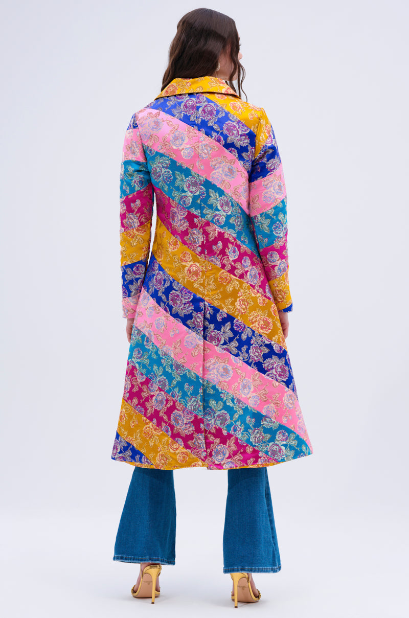 A woman with long brown hair faces away, wearing the BIASED PATCHWORK BROCADE TRENCH—a vibrant diagonally-striped floral coat—paired with blue flared jeans and yellow heels against a plain light background.