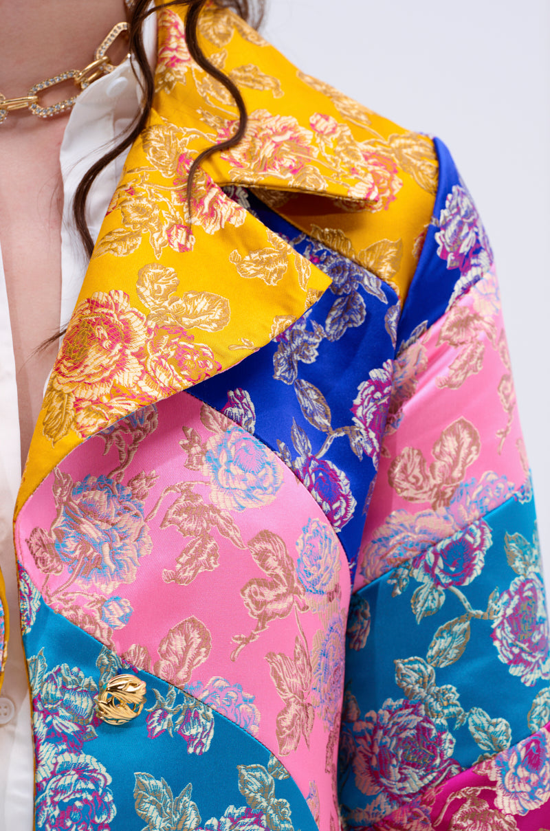 A woman with long brown hair wears the BIASED PATCHWORK BROCADE TRENCH—a vibrant statement coat in yellow, blue, pink, and teal brocade—styled with a gold chain necklace and a white shirt.