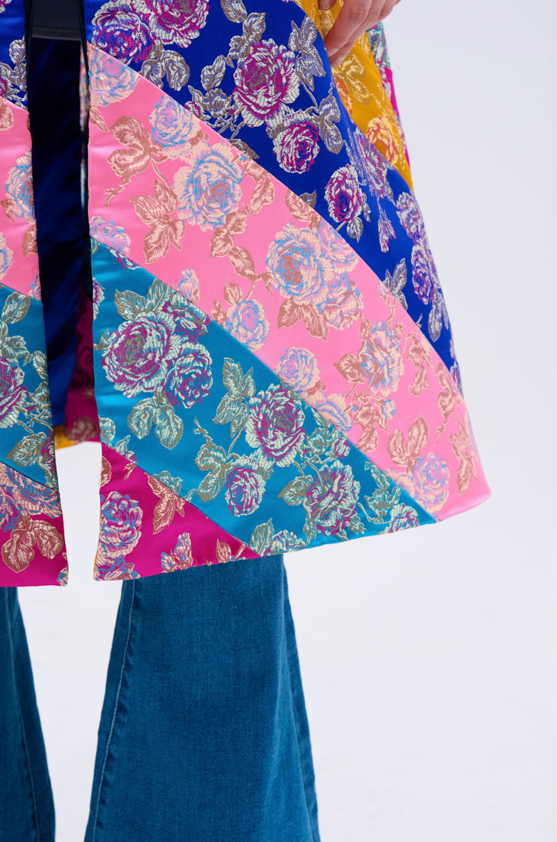 A person wearing blue flared jeans and the BIASED PATCHWORK BROCADE TRENCH—a colorful, floral brocade coat with pink, blue, and yellow diagonal stripes. Only the lower part of the outfit is visible.