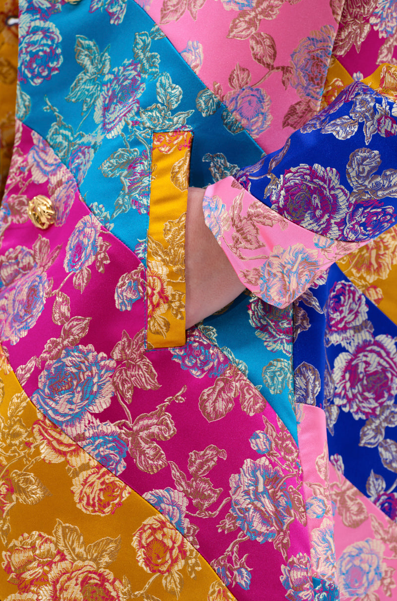 Close-up of the BIASED PATCHWORK BROCADE TRENCH, showcasing vibrant pink, blue, gold, and turquoise floral brocade panels with metallic flower designs and a gold button detail near the pocket.