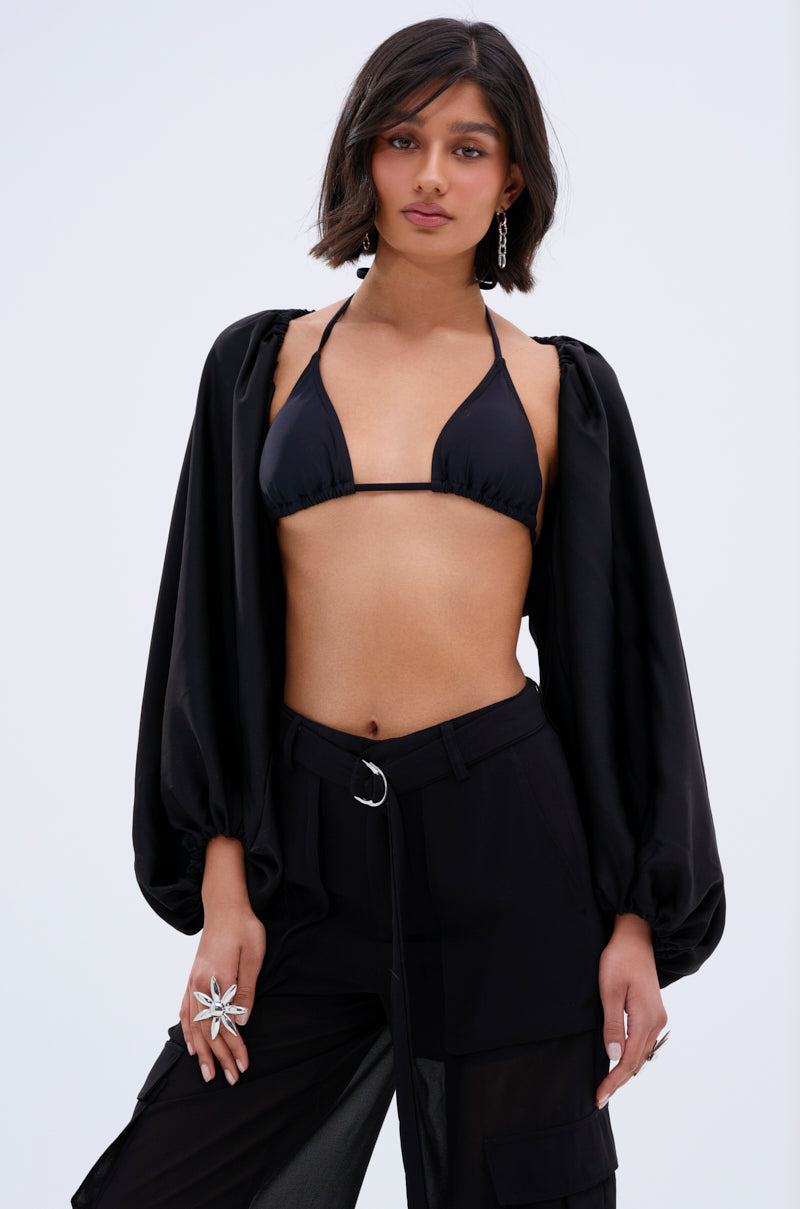 A woman with a short bob, silver earrings, and statement rings poses confidently in a black bikini top, COVER UP CHIFFON SHRUG IN BLACK, and black pants against a plain light background.