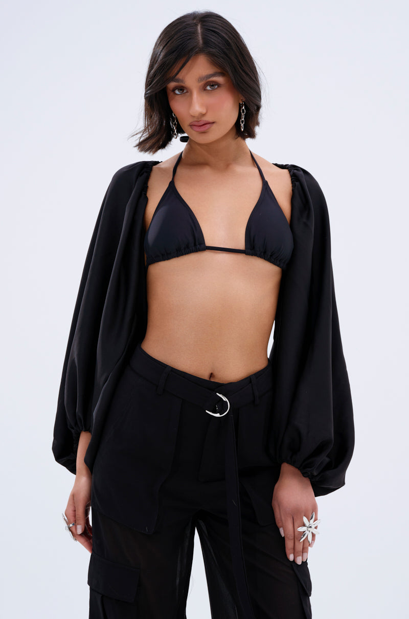 A woman with short dark hair wears a black bikini top, high-waisted pants with a belt, statement ring, earrings, and the COVER UP CHIFFON SHRUG IN BLACK with balloon sleeves, posing confidently against a plain light background.