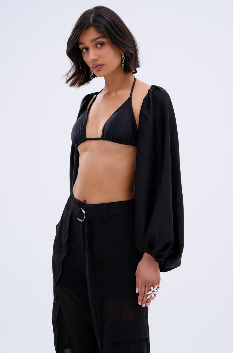 A woman with a bob haircut wears the COVER UP CHIFFON SHRUG IN BLACK over a black bikini top and high-waisted belted pants, paired with statement earrings and a large ring, posing against a plain light background.