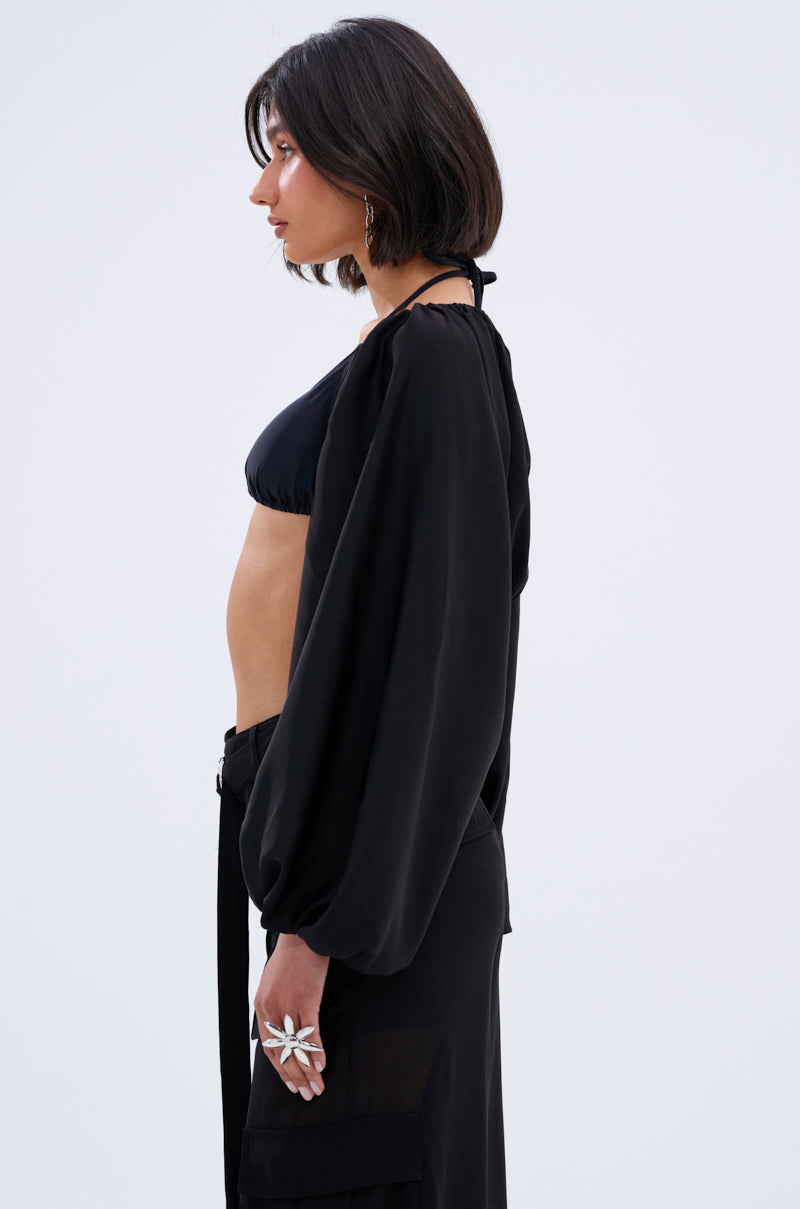 A woman with short dark hair is shown in profile wearing a black halter bikini top and the COVER UP CHIFFON SHRUG IN BLACK, accessorized with a large floral ring against a plain light gray background.