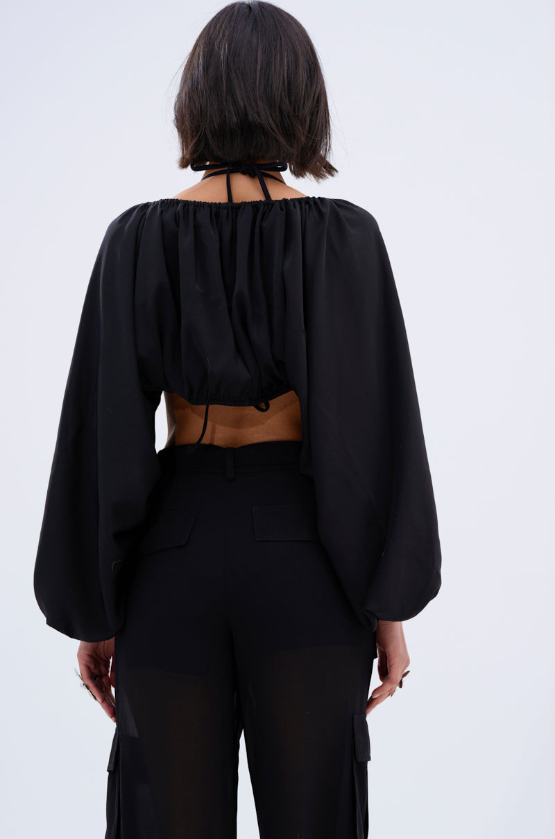 A person with short dark hair is shown from behind, wearing the COVER UP CHIFFON SHRUG IN BLACK over high-waisted black pants against a plain light background.